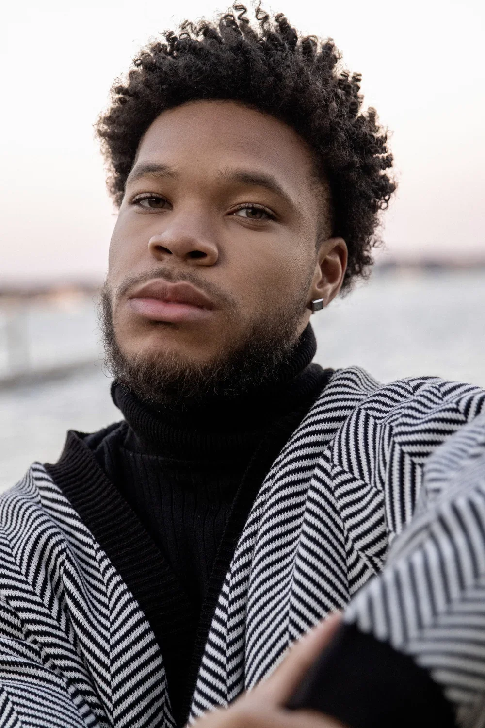 A young man with curly hair and facial hair taking a selfie outdoors, wearing a striped jacket over a black turtleneck, with water and blurred landscape in the background.