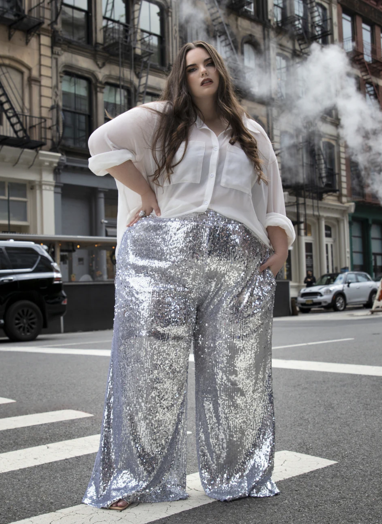 A woman standing on a city street crosswalk, wearing a white blouse and shiny silver sequin wide-legged pants, with buildings and smoke in the background.