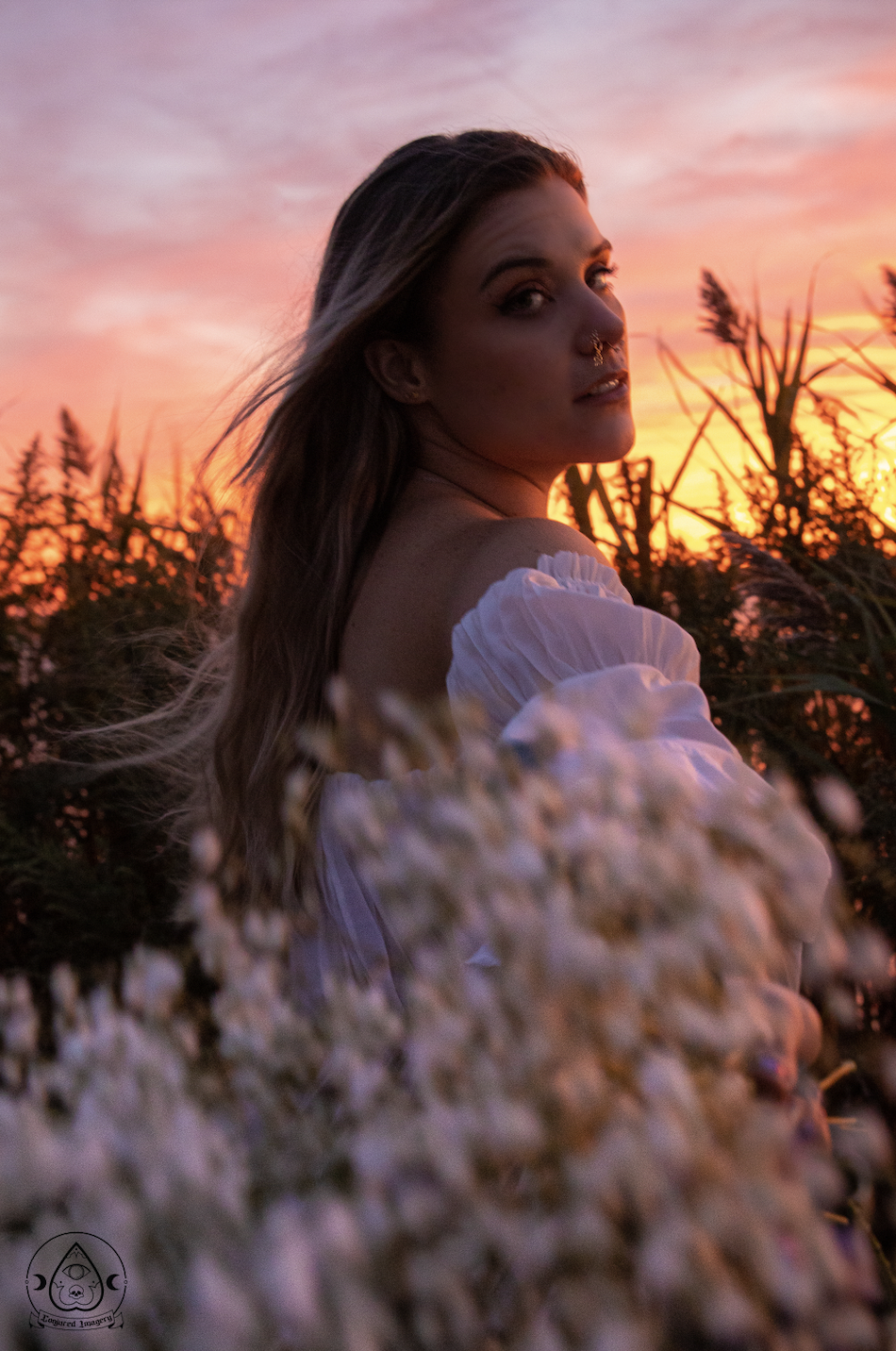 A woman with long hair and a nose piercing, wearing a white off-shoulder top, standing in a field of tall plants or flowers during a colorful sunset.