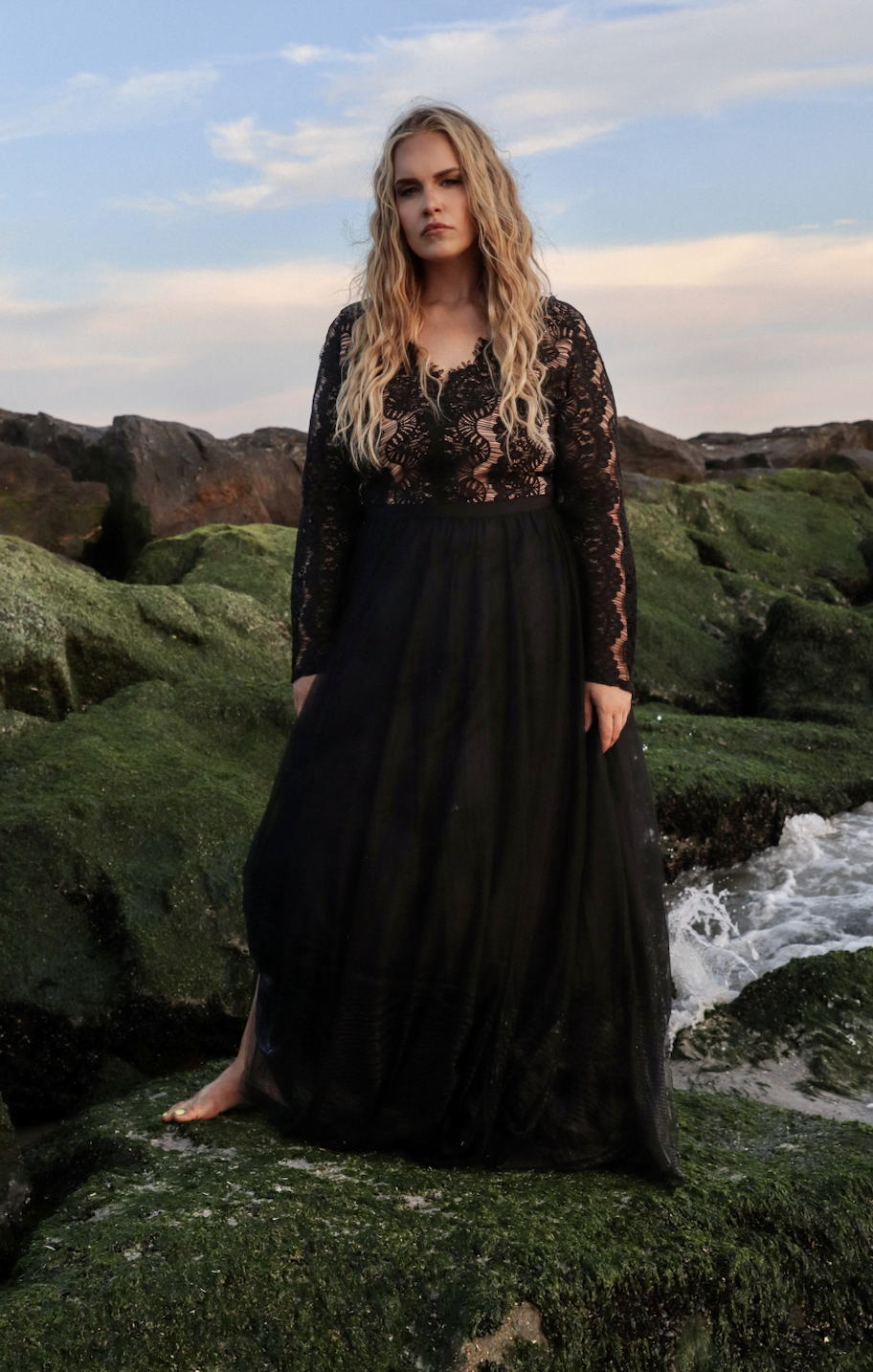 A woman in a black lace dress standing barefoot on green moss-covered rocks by the water at sunset.