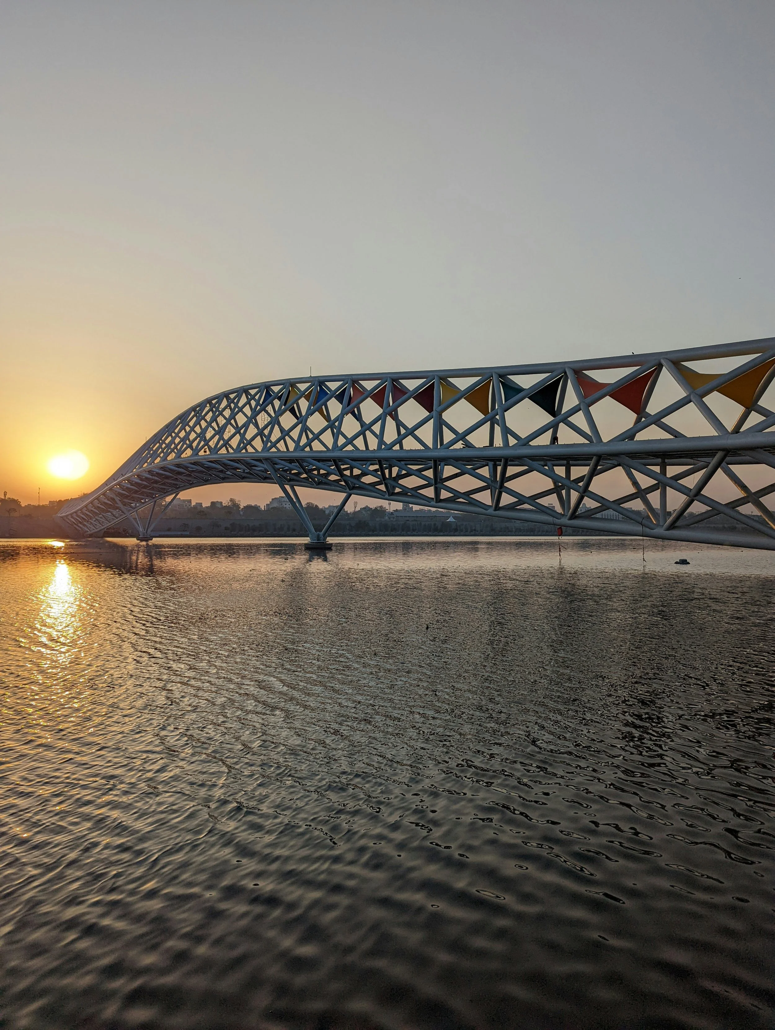 A modern white footbridge with a lattice structure arching over a body of water at sunset.