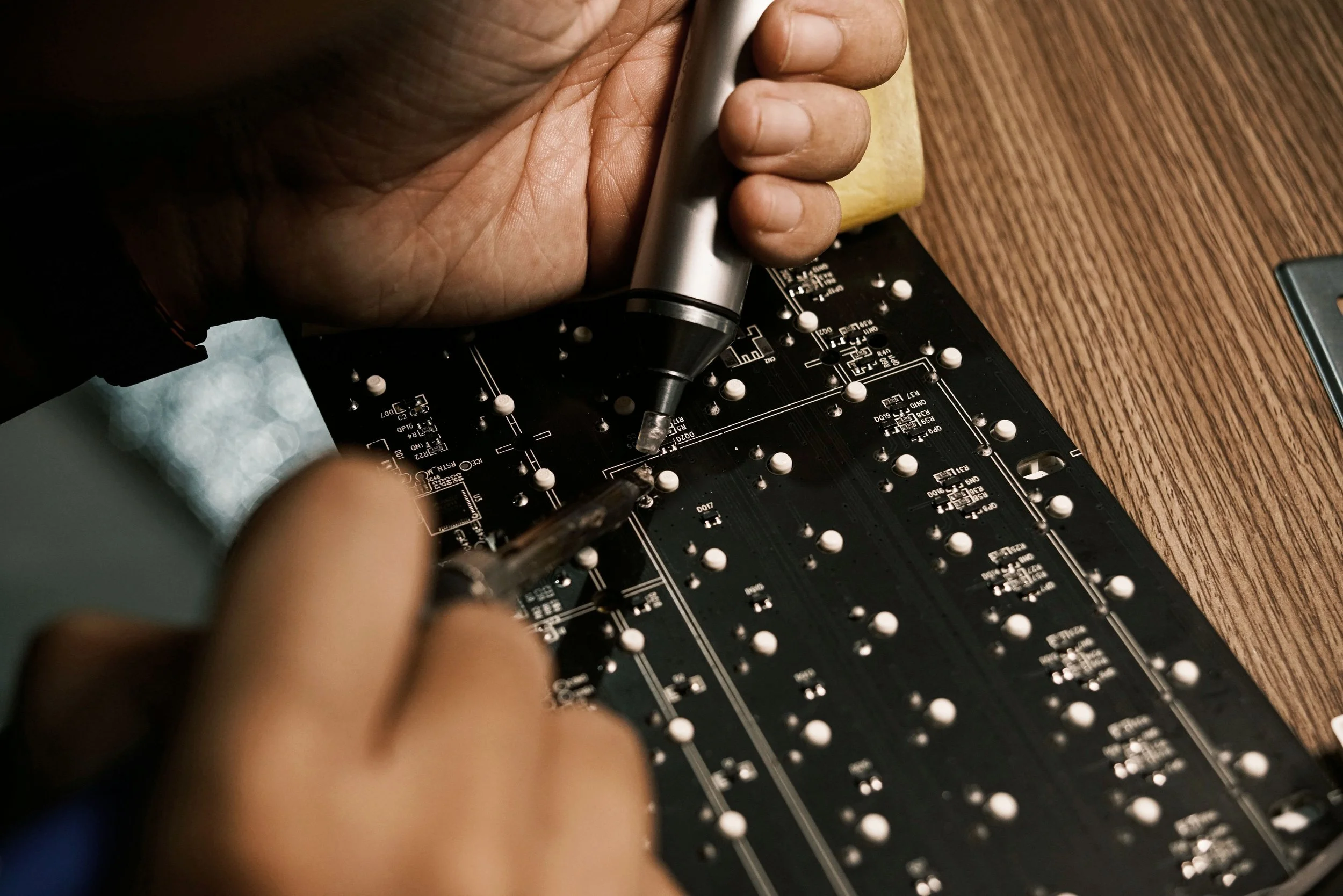 Hands working on assembling a black printed circuit board with small white components using a soldering iron.