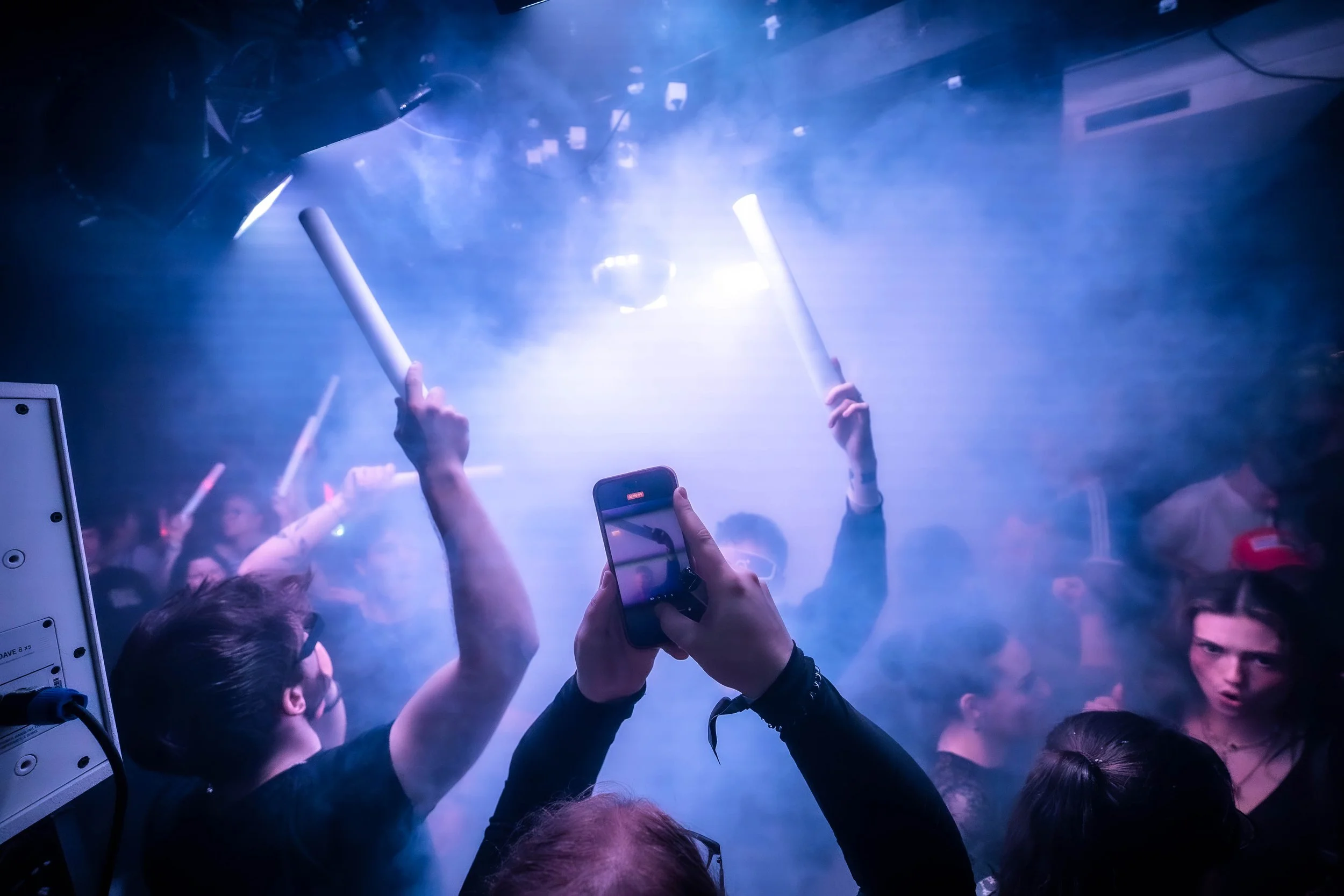 Menschen feiern in einer dunklen, rauchigen Umgebung, einige halten leuchtende Gegenstände in den Händen, und eine Person benutzt ihr Smartphone, um Bilder oder Videos aufzunehmen.