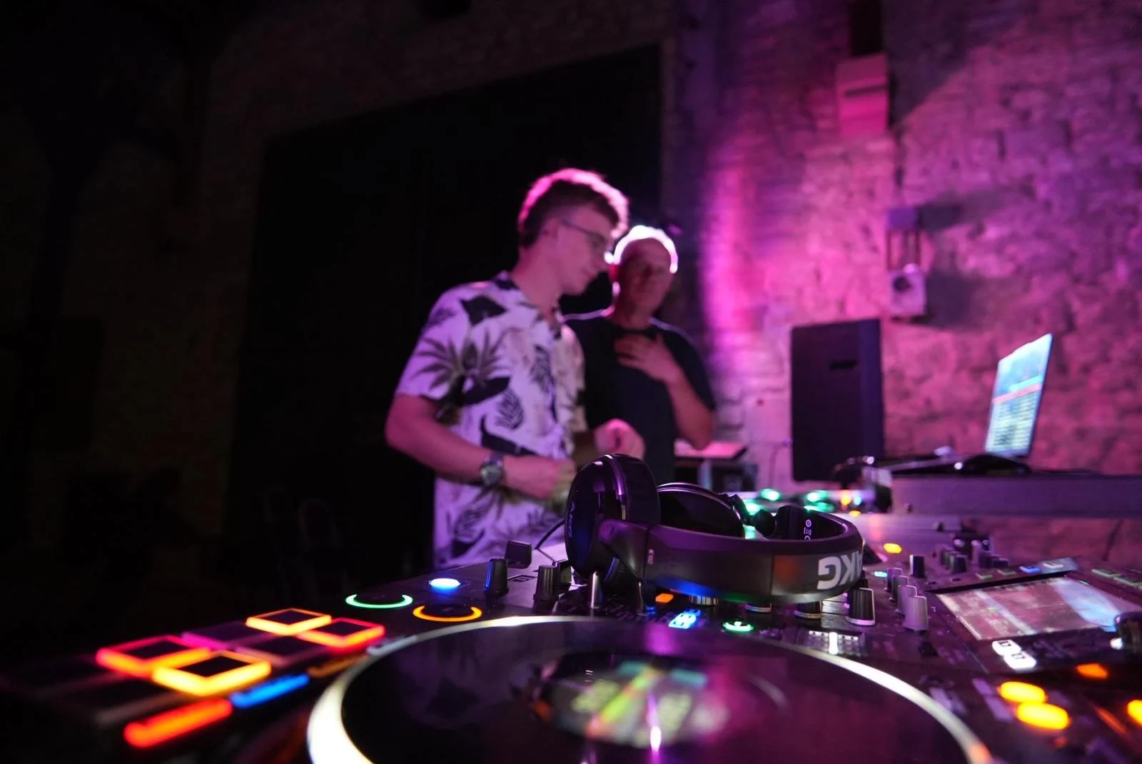 Zwei DJs stehen an einem DJ-Setup mit bunten Lichtern, in einer dunklen, brick-wände Umgebung, beim Auflegen.