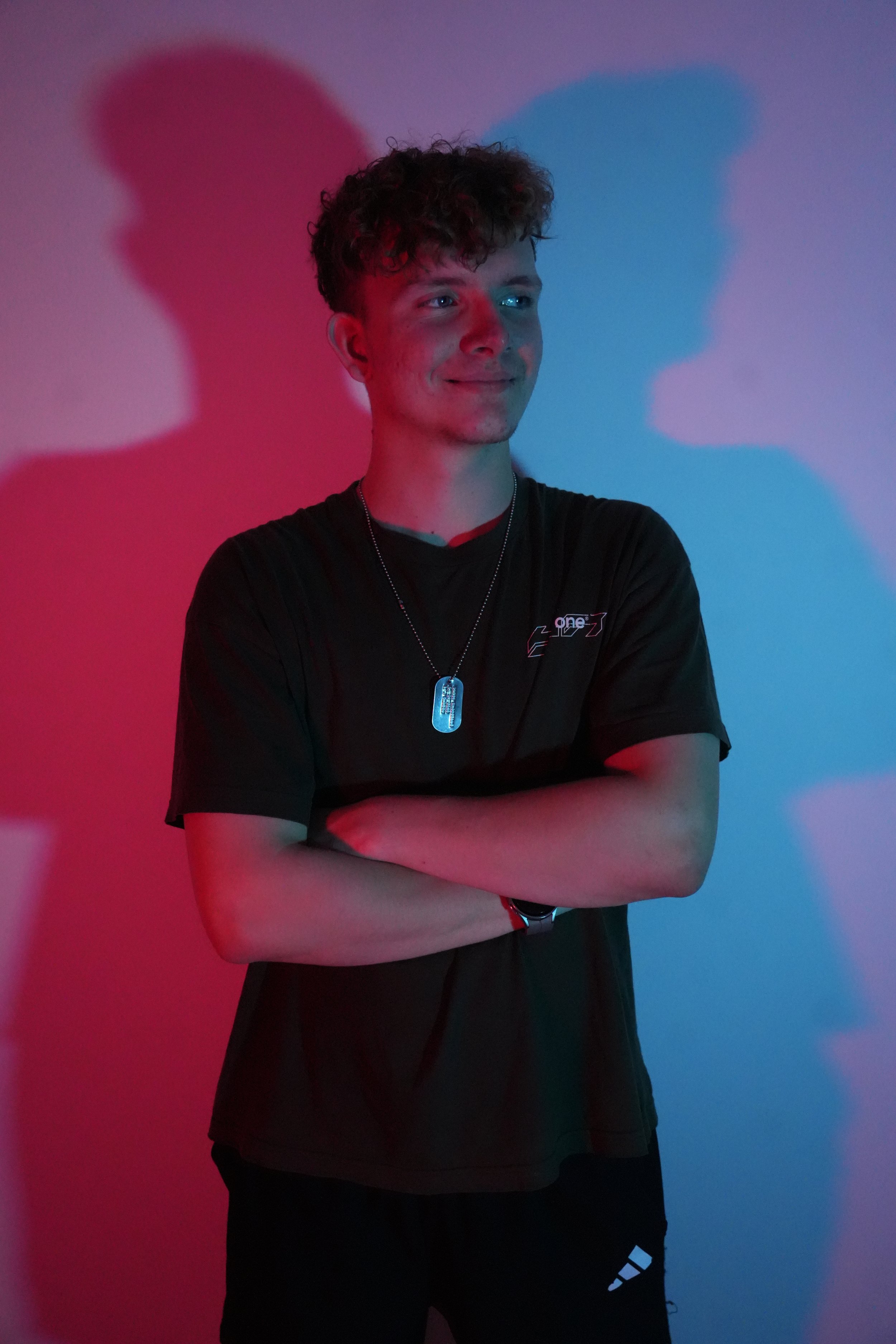 DJ - Dybaxxz & Geschäftsführer Paul-J. Zander
Junger Mann mit kurzen, lockigen Haaren trägt schwarze Sportkleidung und ein Dogtag-Halsband, steht vor einer Wand mit roten und blauen Schatten, die sich im Licht widerspiegeln.