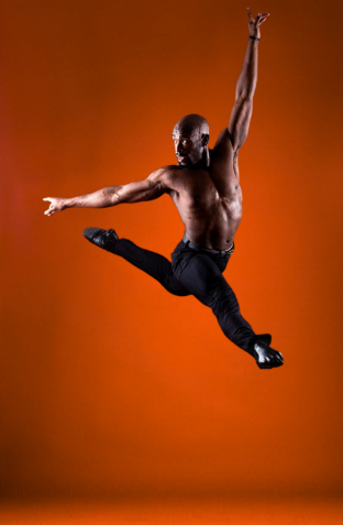 A shirtless man with dark skin performing a jump with one arm raised and one arm extended to the side against an orange background.