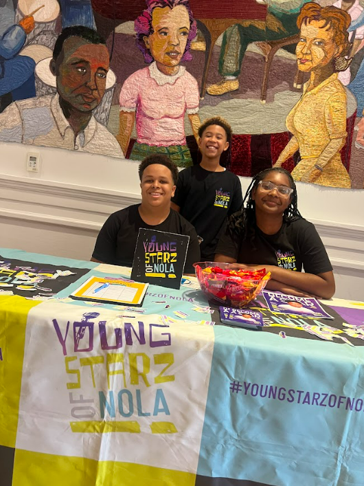 Three smiling young children sitting and standing behind a table at a Young Starz of NOLA event, with a large colorful mural of diverse people painted on the wall behind them.