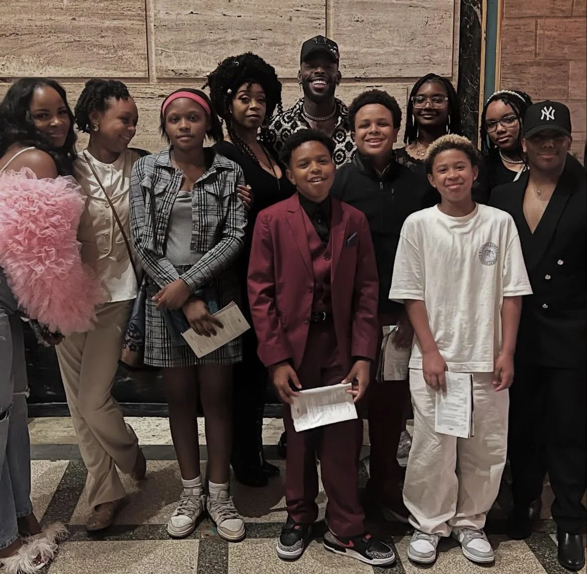 Group portrait of twelve children and two adults standing together indoors, dressed in a mix of casual and formal attire, smiling at the camera.