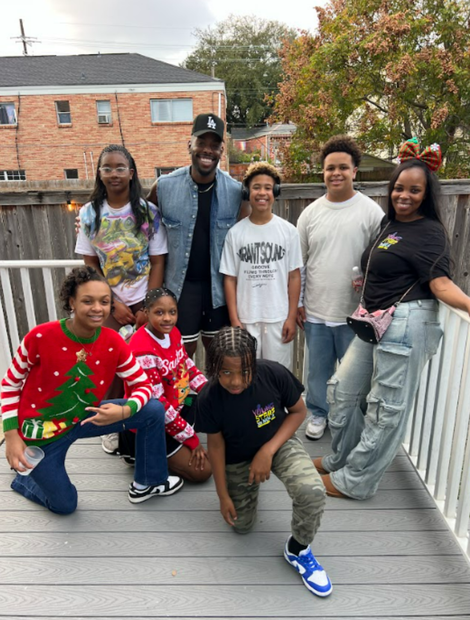 Group of children and adults posing on a wooden deck, with some wearing festive Christmas sweaters, in a backyard with a tree and brick houses in the background.