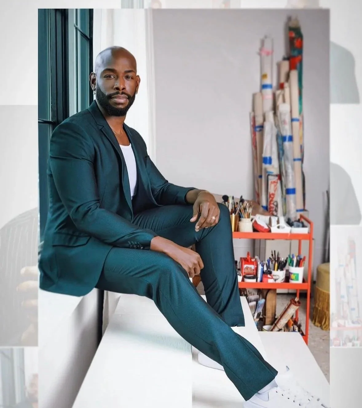 A man in a black suit sitting on a white surface in an art studio, with art supplies and rolled posters in the background.