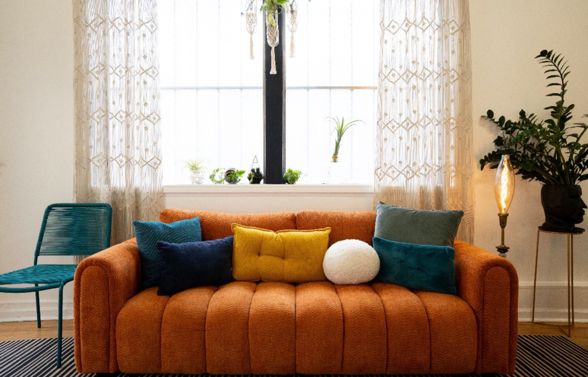 A cozy living room with an orange couch decorated with colorful pillows, a blue chair to the left, a window with sheer beige curtains in the background, and a tall potted plant with dark green leaves on the right next to a vintage floor lamp, with a collection of small potted plants on the windowsill.