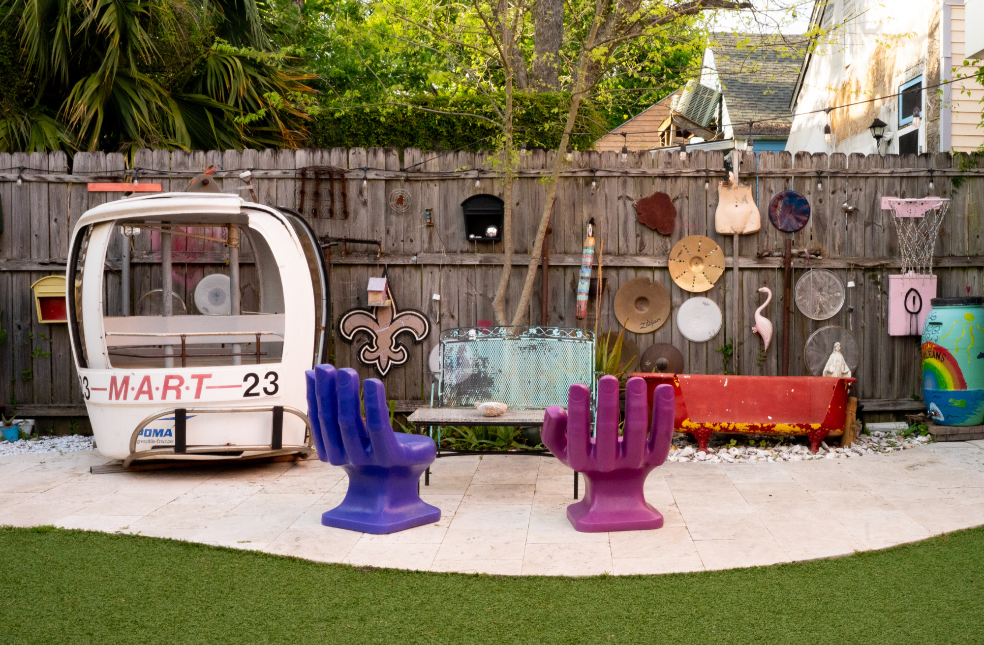 A backyard with a wooden fence decorated with various artistic objects and wall hangings, a white gondola-like structure, two purple and pink sculptural chairs shaped like hands, a metal table with a large stone on top, a red clawfoot bathtub, a large painted rain barrel, and an artificial grass lawn in the foreground.