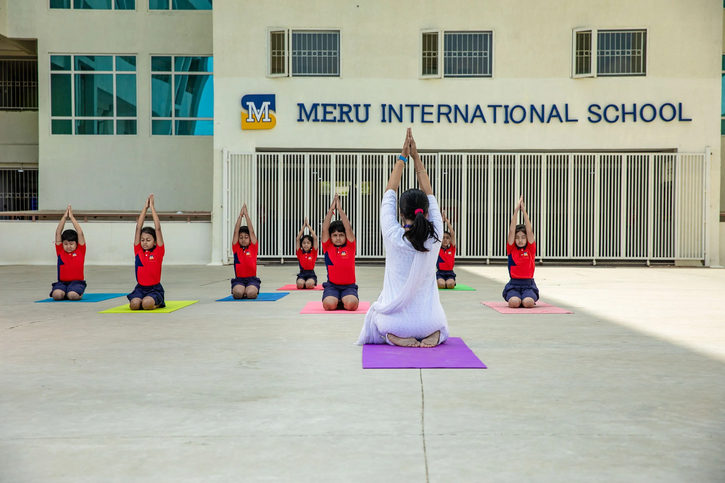 A group of students in red and white uniforms practicing yoga on purple mats in an outdoor courtyard, with the Meru International School building in the background.