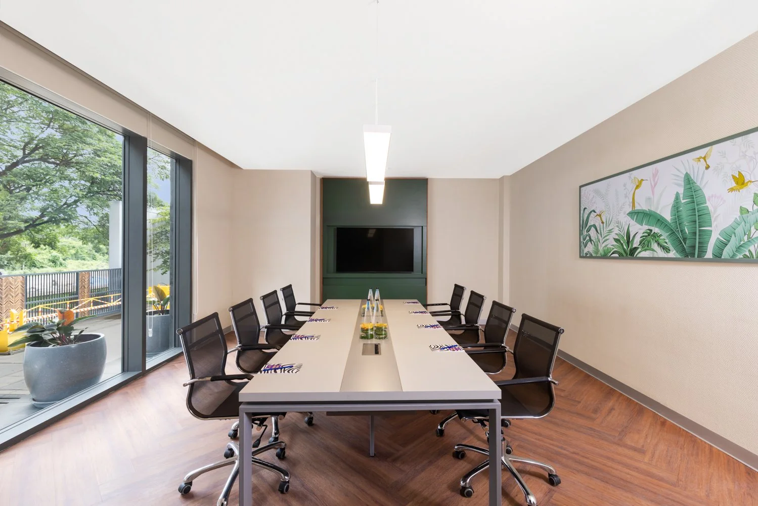 nterior photography of a brightly lit hotel boardroom featuring a long meeting table, black office chairs, a wall-mounted TV, and wooden floors