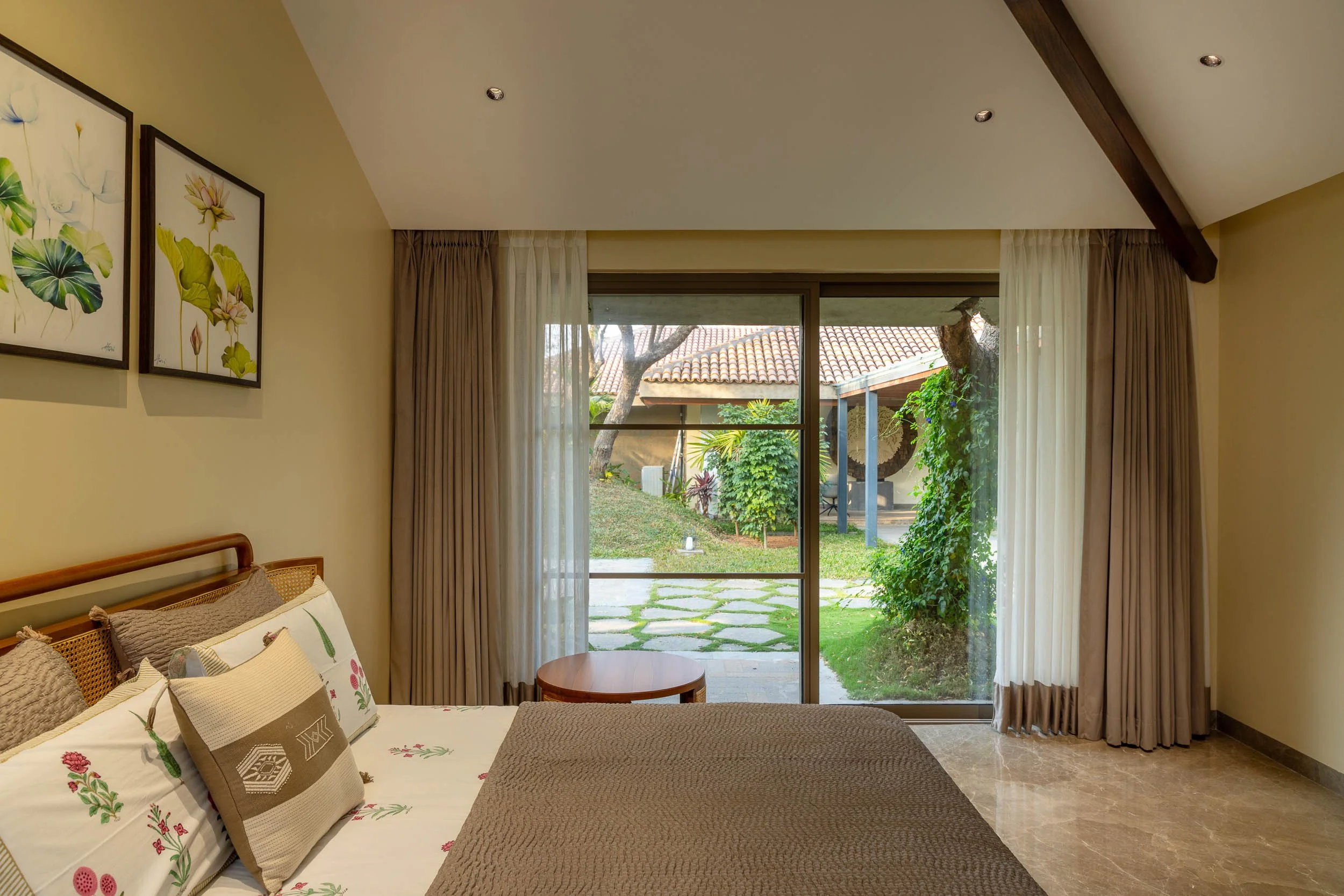 Interior photography of a luxury resort farm stay bedroom featuring a rustic wooden ceiling beam, a large bed, and glass sliding doors opening to a green garden pathway.
