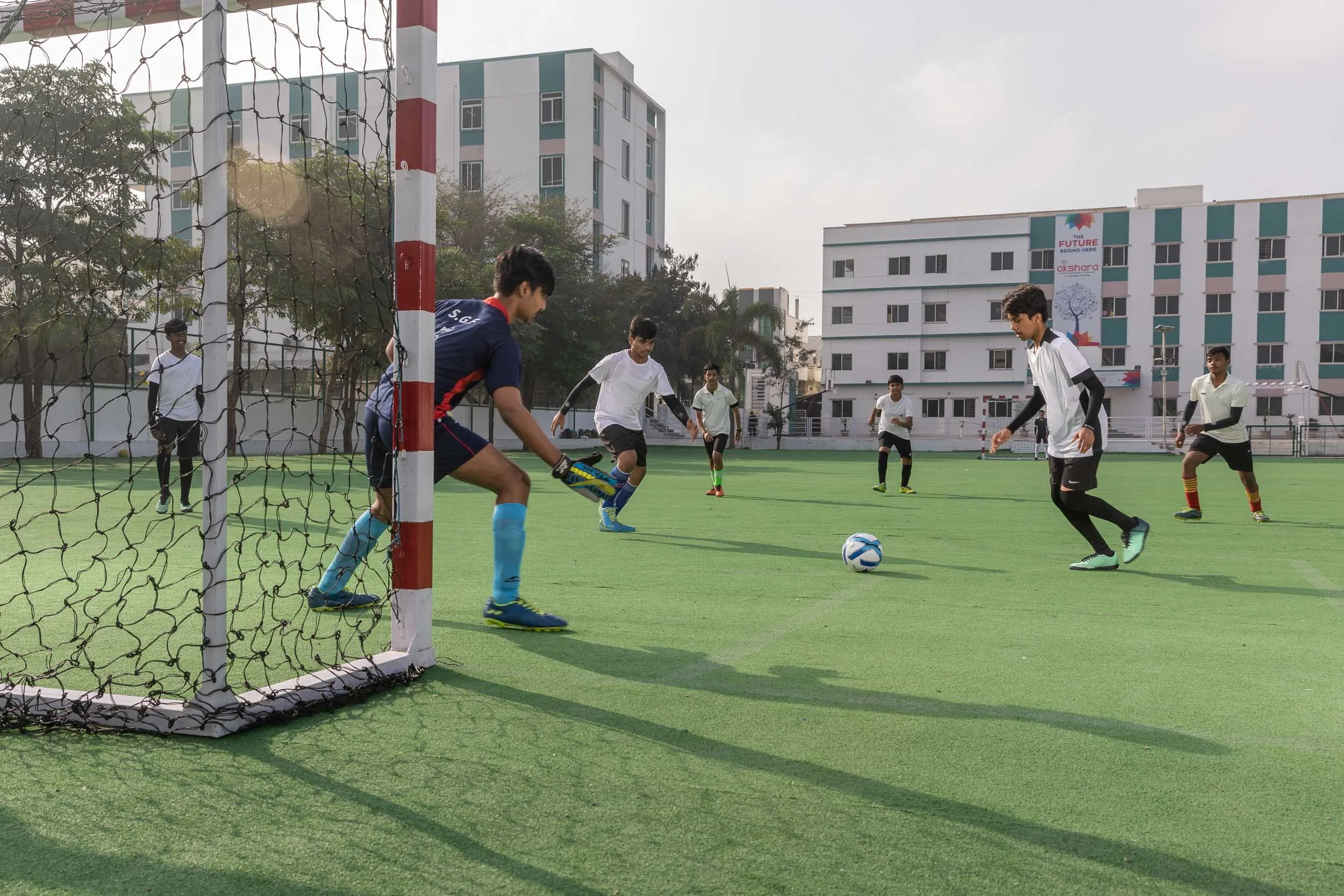 A group of male students in sports uniforms playing football on a bright green artificial turf field. One student is kicking the ball toward a goal net, with large modern school buildings in the background.