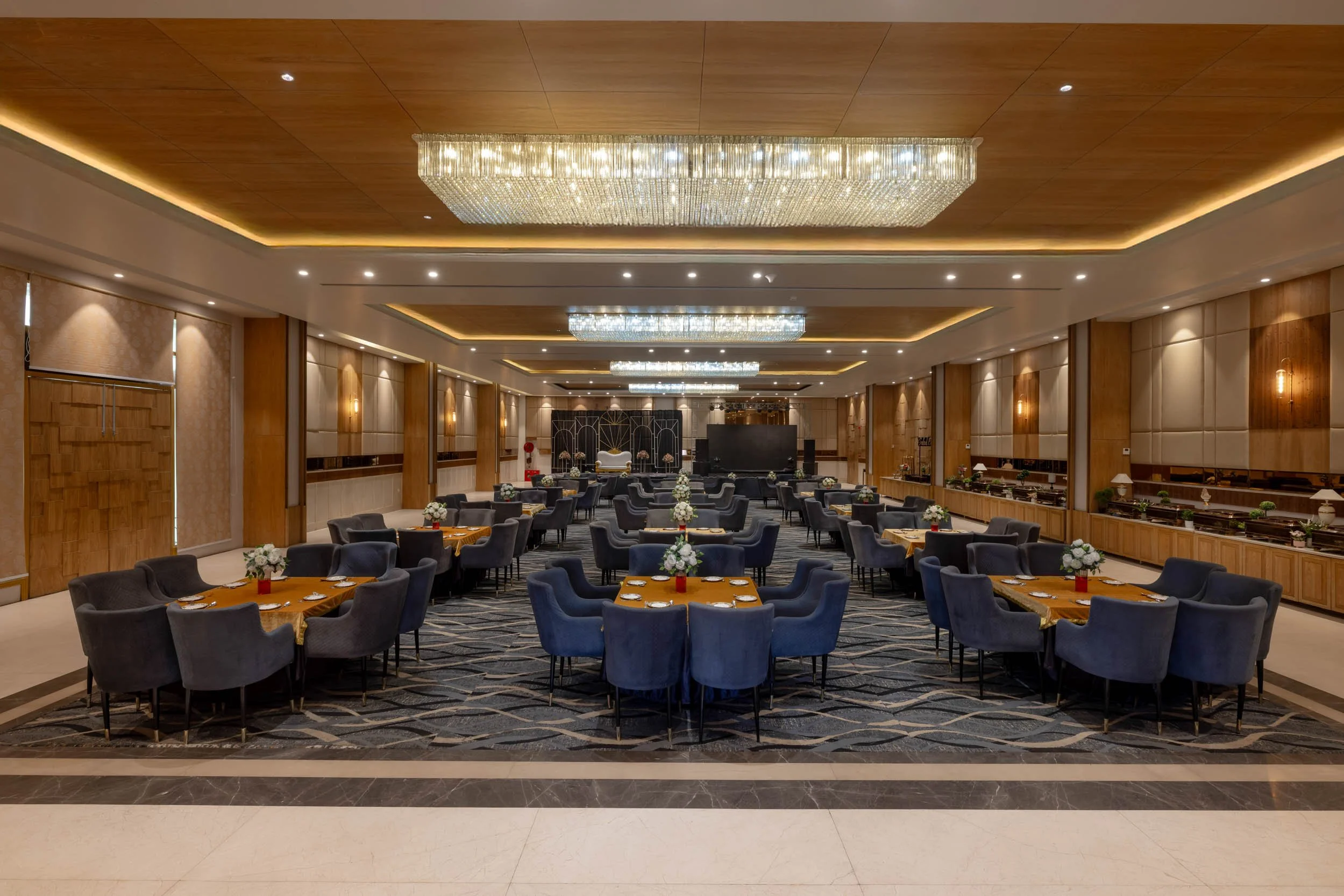 Interior photography of a luxury hotel banquet hall featuring round dining tables, blue upholstered chairs, patterned carpet, and a large central chandelier