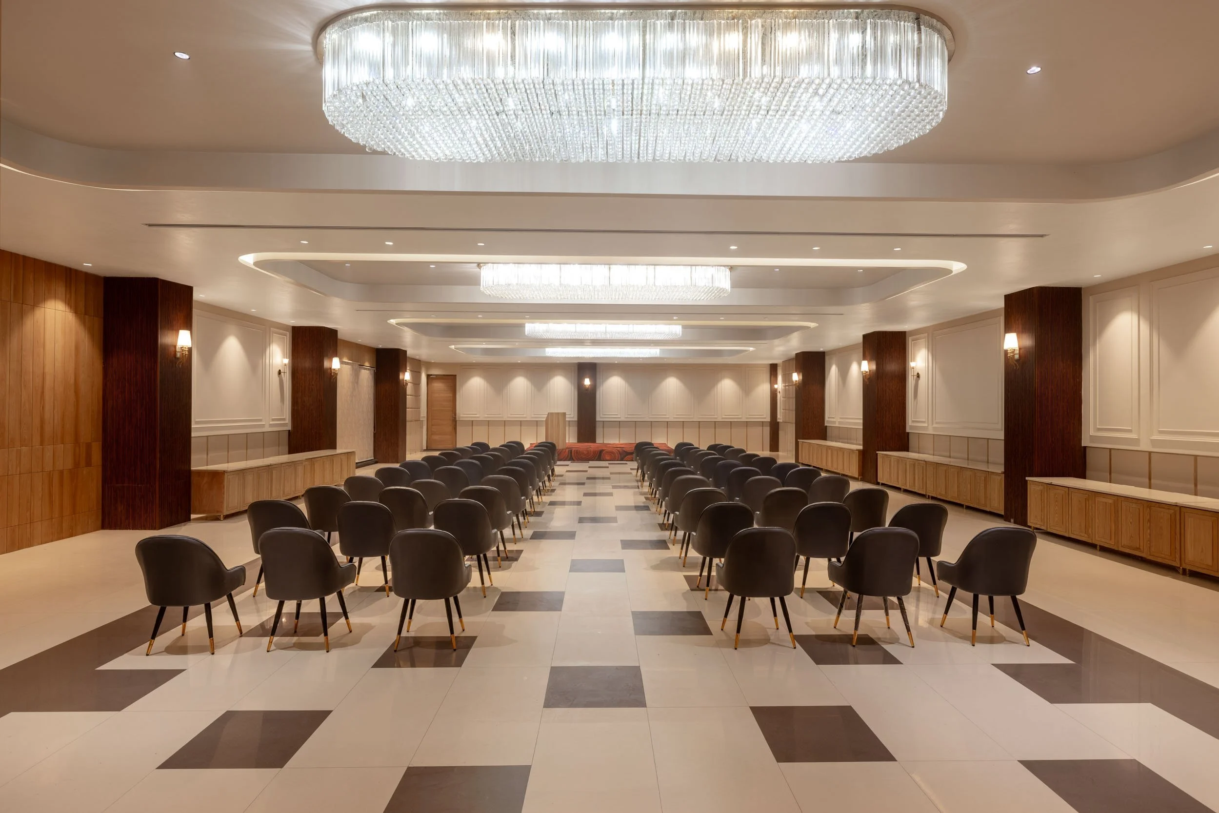 Interior photography of a luxury hotel conference hall featuring rows of dark chairs, patterned flooring, wood-paneled walls, and a large central crystal chandelier