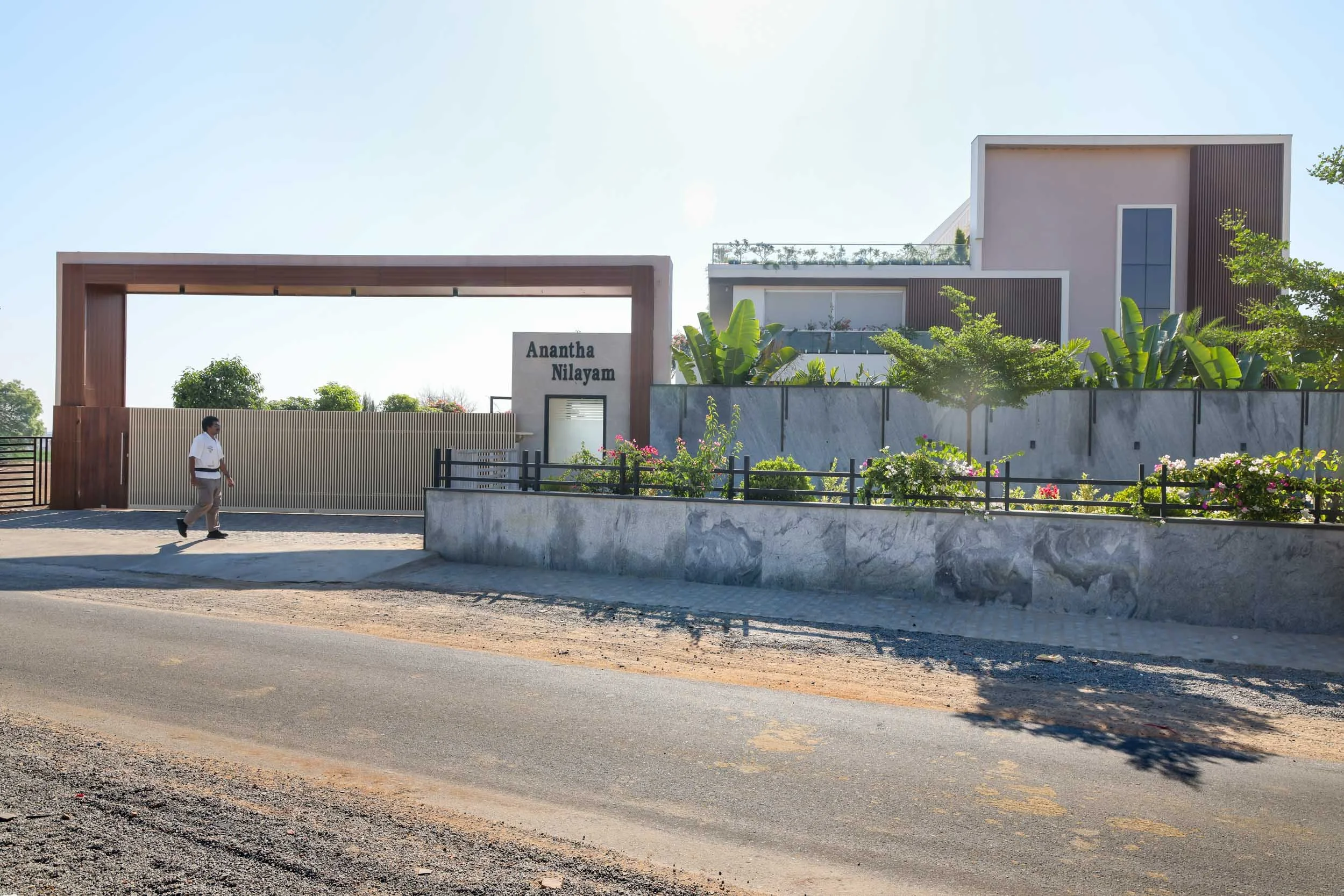Professional exterior architectural photography of a sprawling modern residential home, capturing a wide street view, a striking entrance gate, and a beautifully landscaped boundary wal