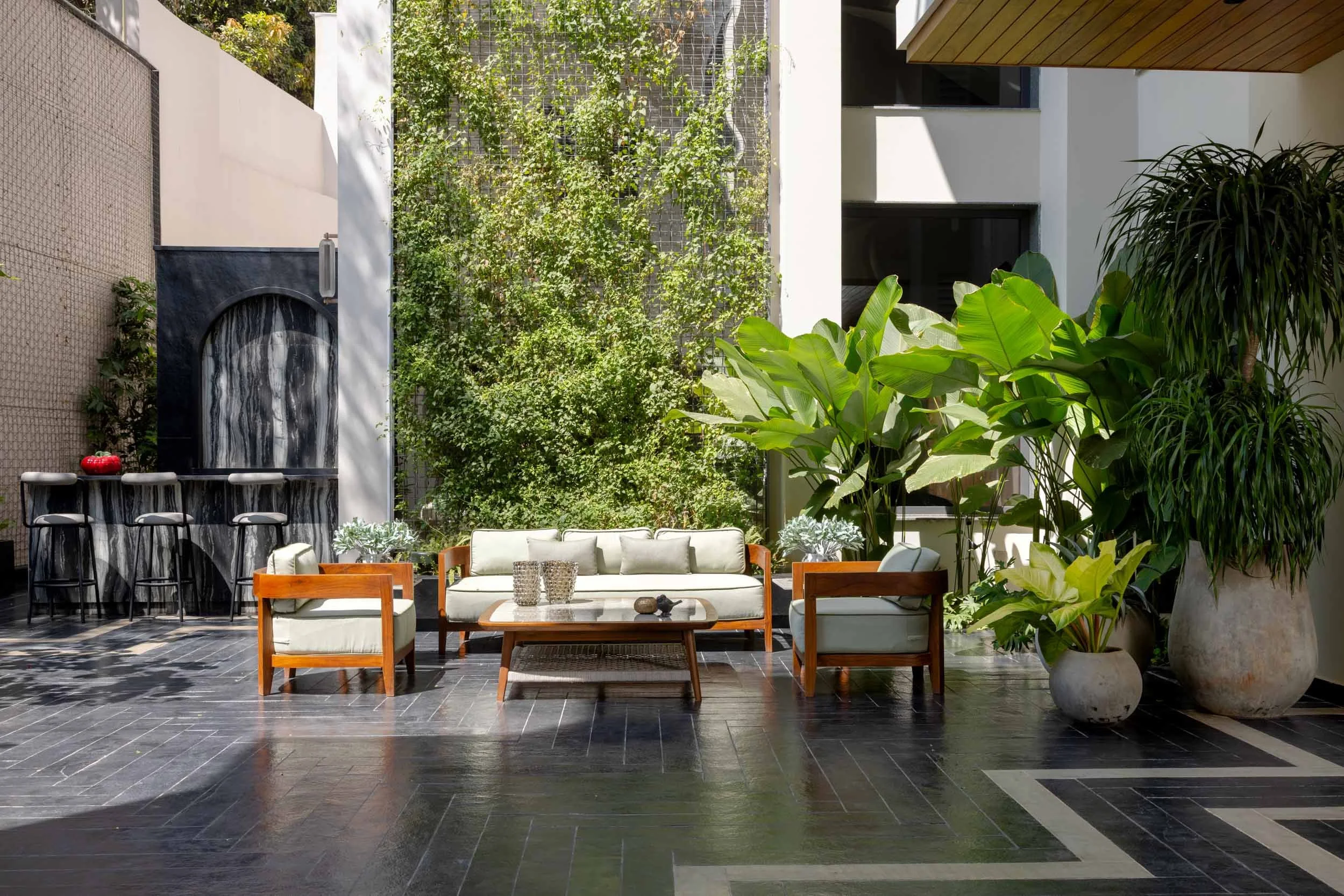 Modern outdoor courtyard patio featuring a wooden seating arrangement on dark reflective stone flooring, surrounded by lush tropical plants