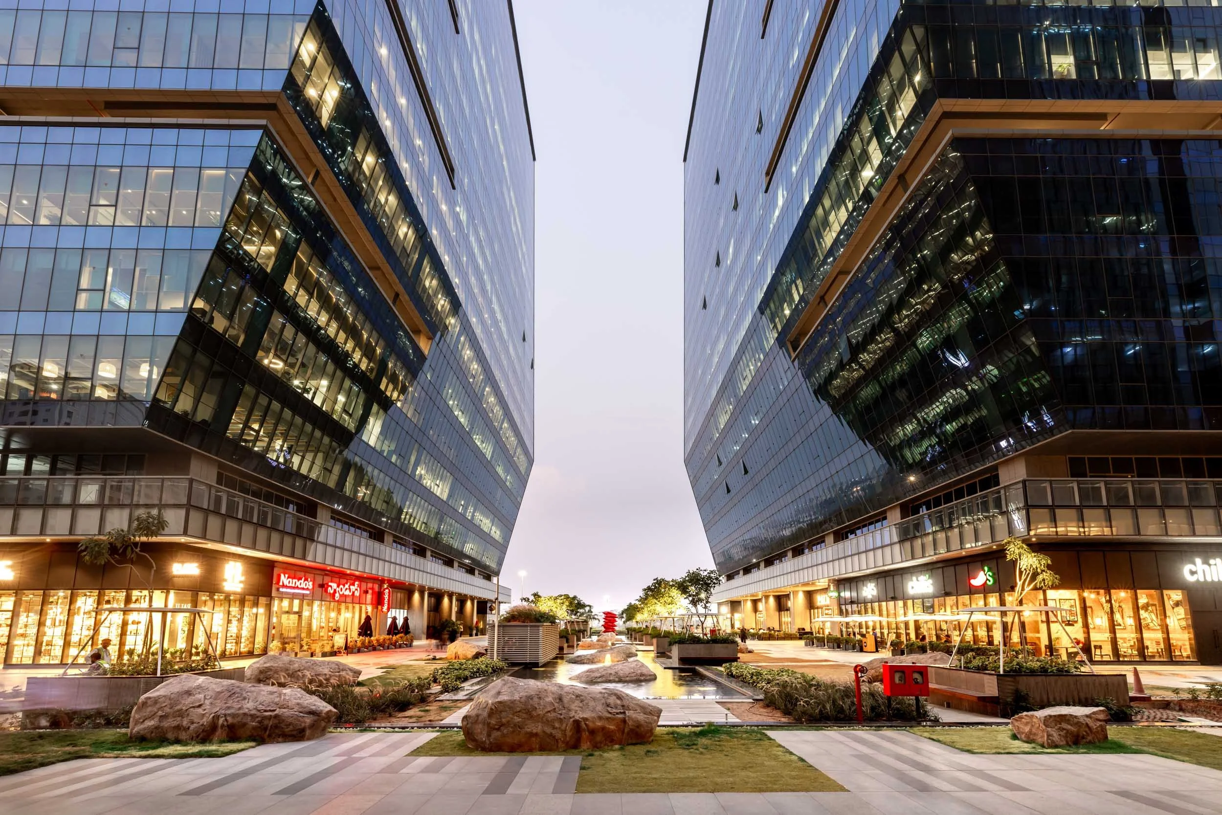 Professional commercial architecture photography capturing a dynamic twilight view of a modern business district, highlighting sleek glass facades, warm interior lighting, and a striking street-level perspective.