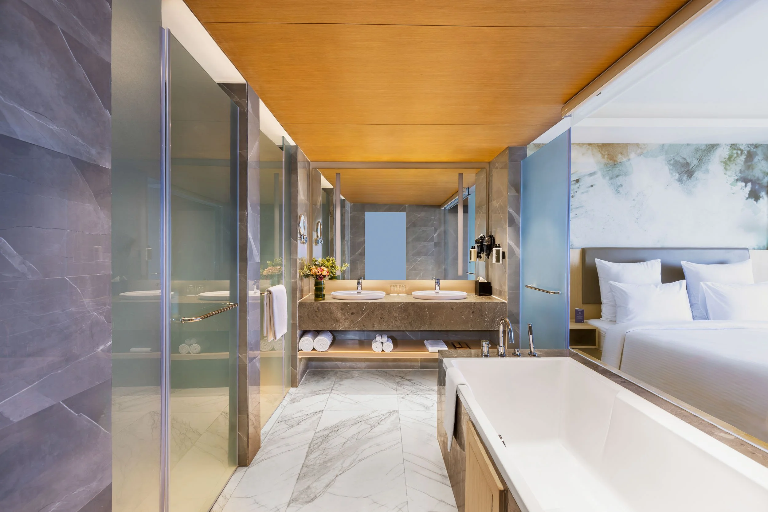 Interior photography of a modern luxury hotel bathroom featuring a long white dual vanity, warm wooden ceiling panels, large mirrors, and a glass shower enclosure