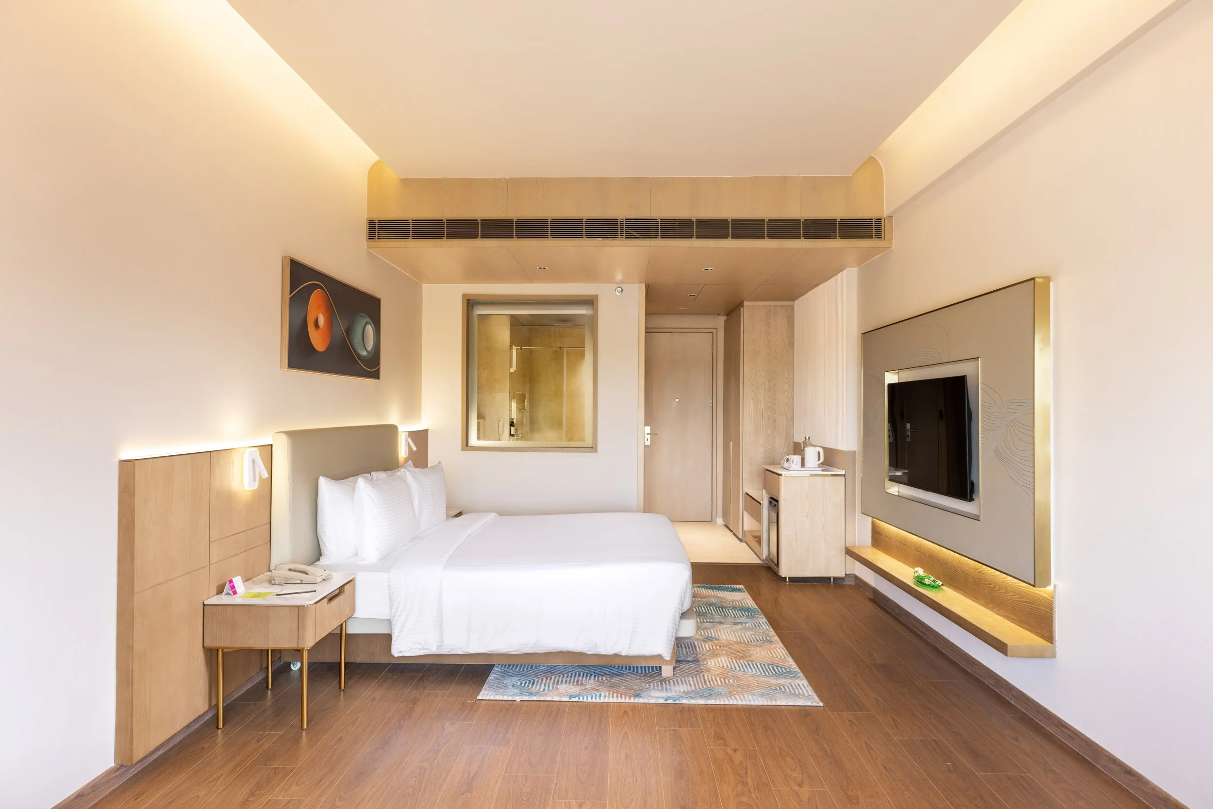 Interior photography of a luxury hotel bedroom featuring a bed with a wood headboard, warm bedside lamps, framed wall art, and a wall-mounted TV.