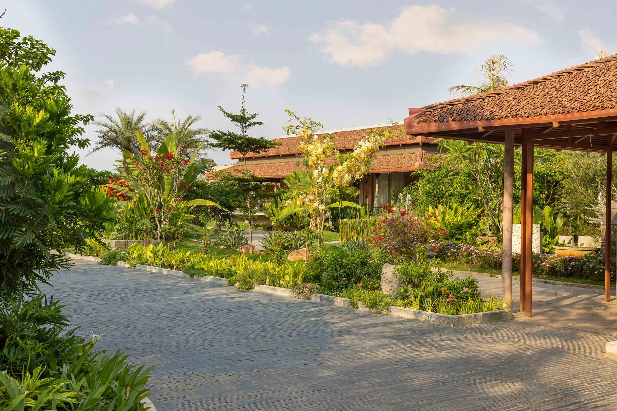 Exterior daytime photography of a luxury resort farm stay featuring a wide stone pathway winding through lush green gardens toward a traditional pitched-roof building