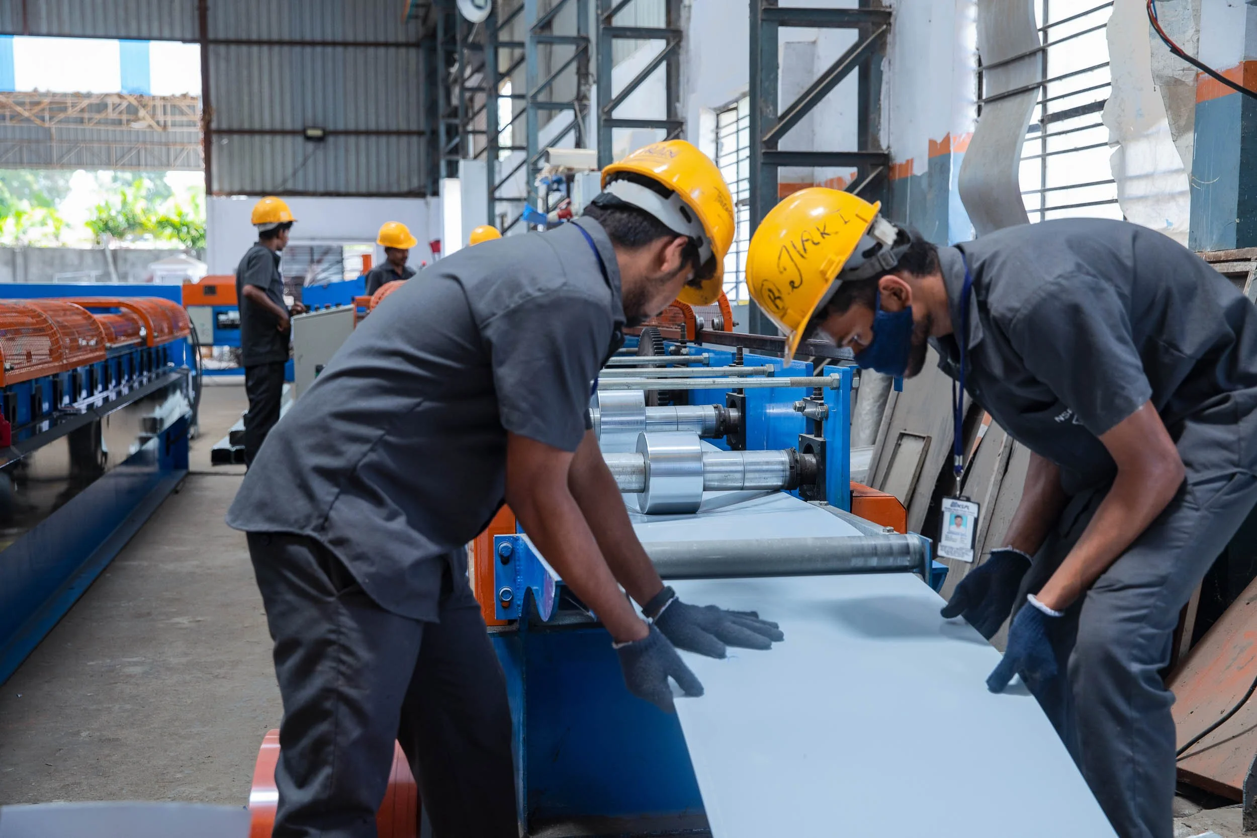 Interior industrial photography of two factory workers in yellow hard hats inspecting a large flat blue sheet of material on the production line in a manufacturing plant