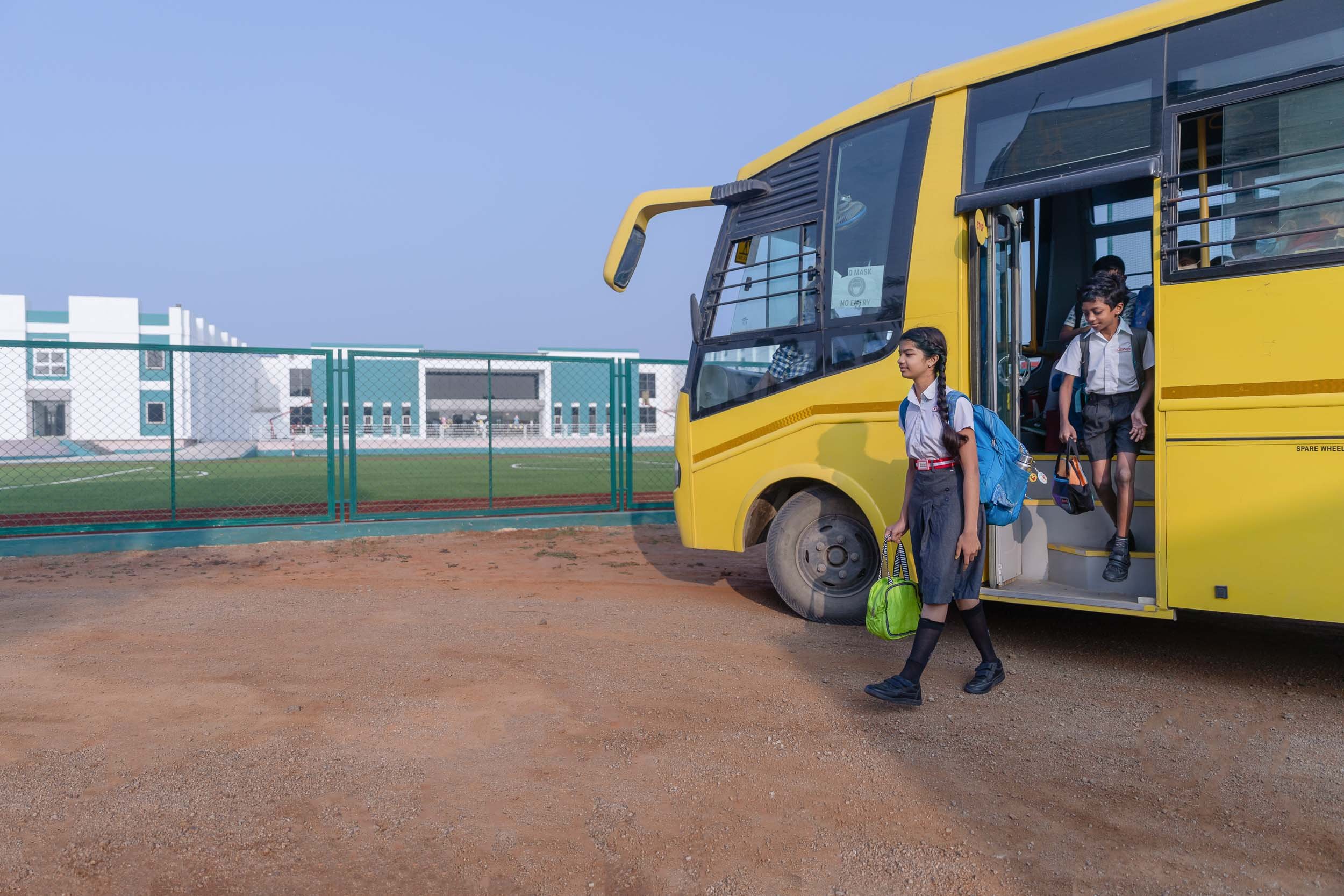 Students in school uniforms standing near the open door of a yellow school bus parked on a campus driveway, with a green fence and school building in the background