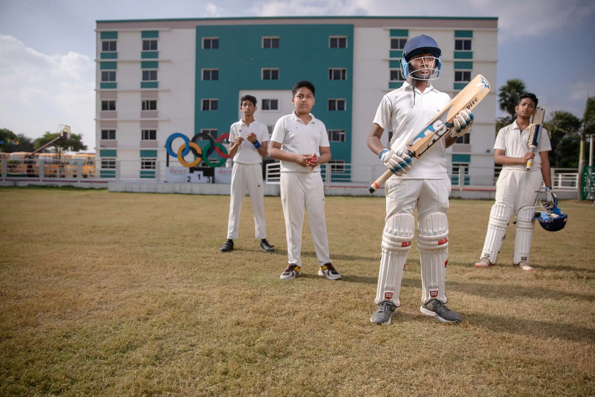 A group of male students in white cricket uniforms standing together on a school sports field. The student in the front is holding a cricket bat, with a large, modern school building visible in the background