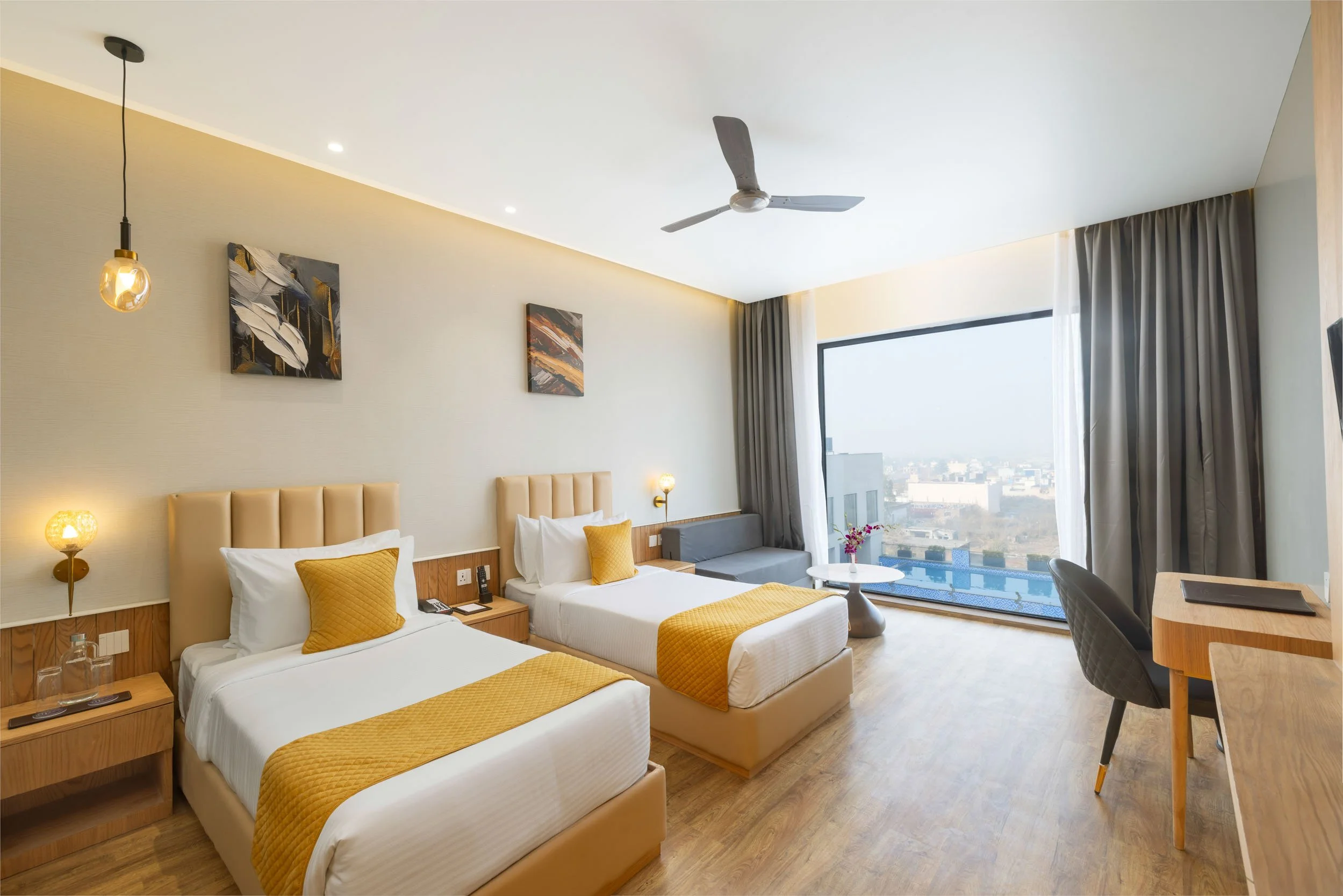 Interior photography of a modern hotel twin room featuring two beds with yellow runners, wooden flooring, a ceiling fan, and a large window with city views
