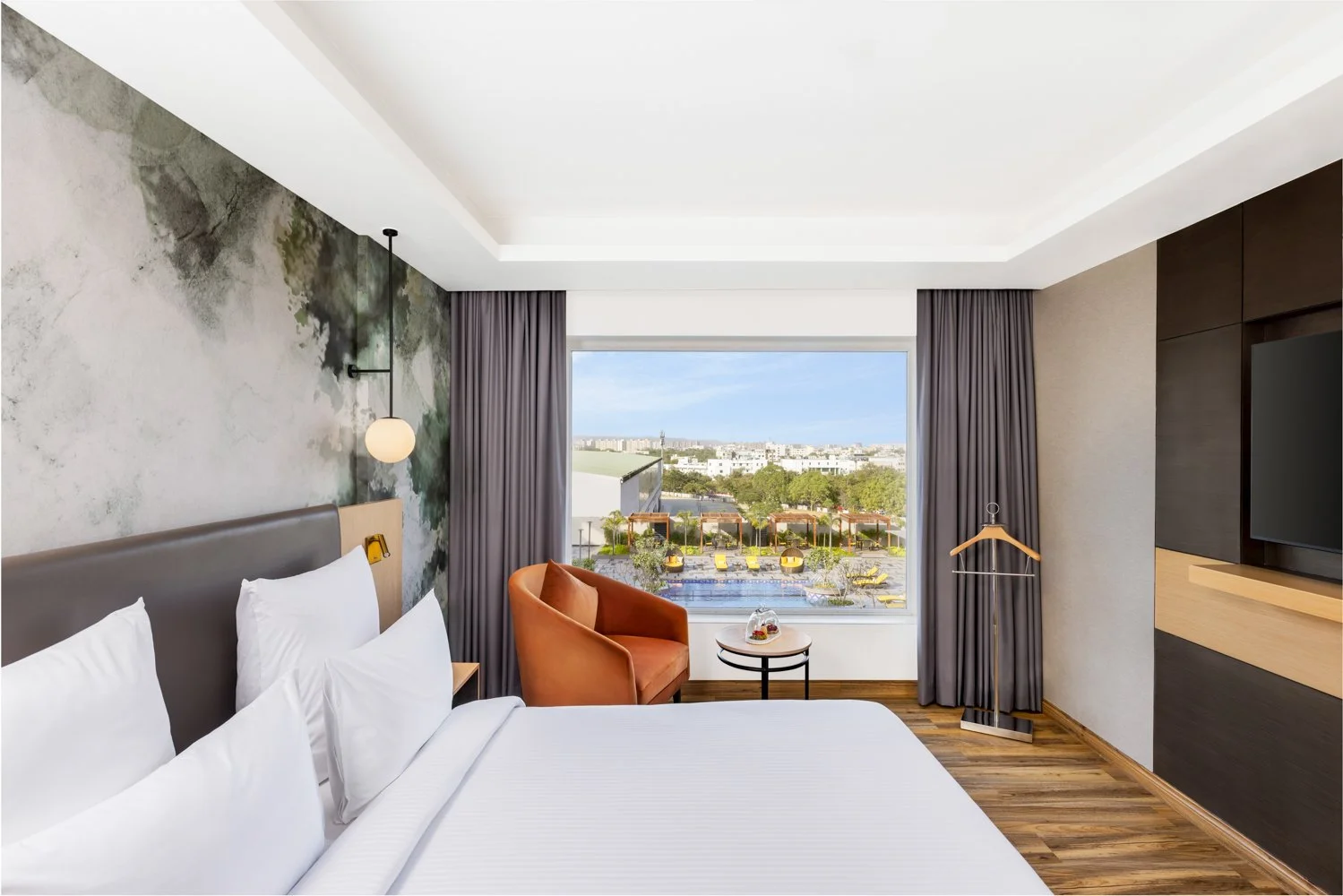 nterior photography of a modern hotel bedroom featuring a nature-inspired accent wall, a bright orange armchair, a large white bed, and a wall-mounted TV
