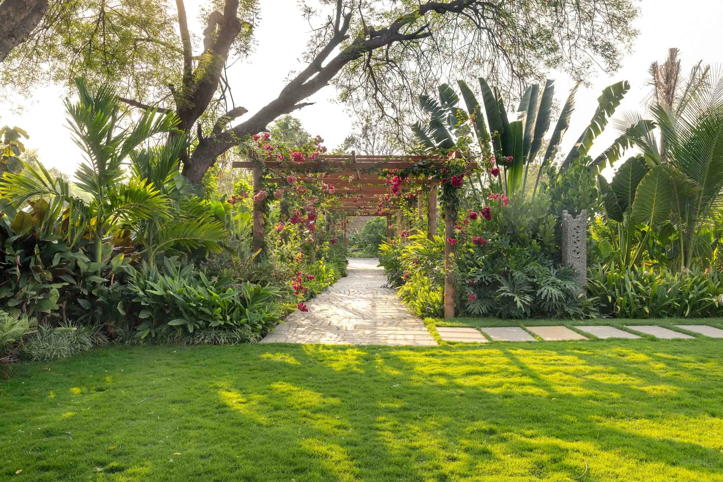 Exterior daytime photography of a luxury resort farm stay featuring a lush green lawn, a stone pathway, tropical plants, and a wooden gazebo