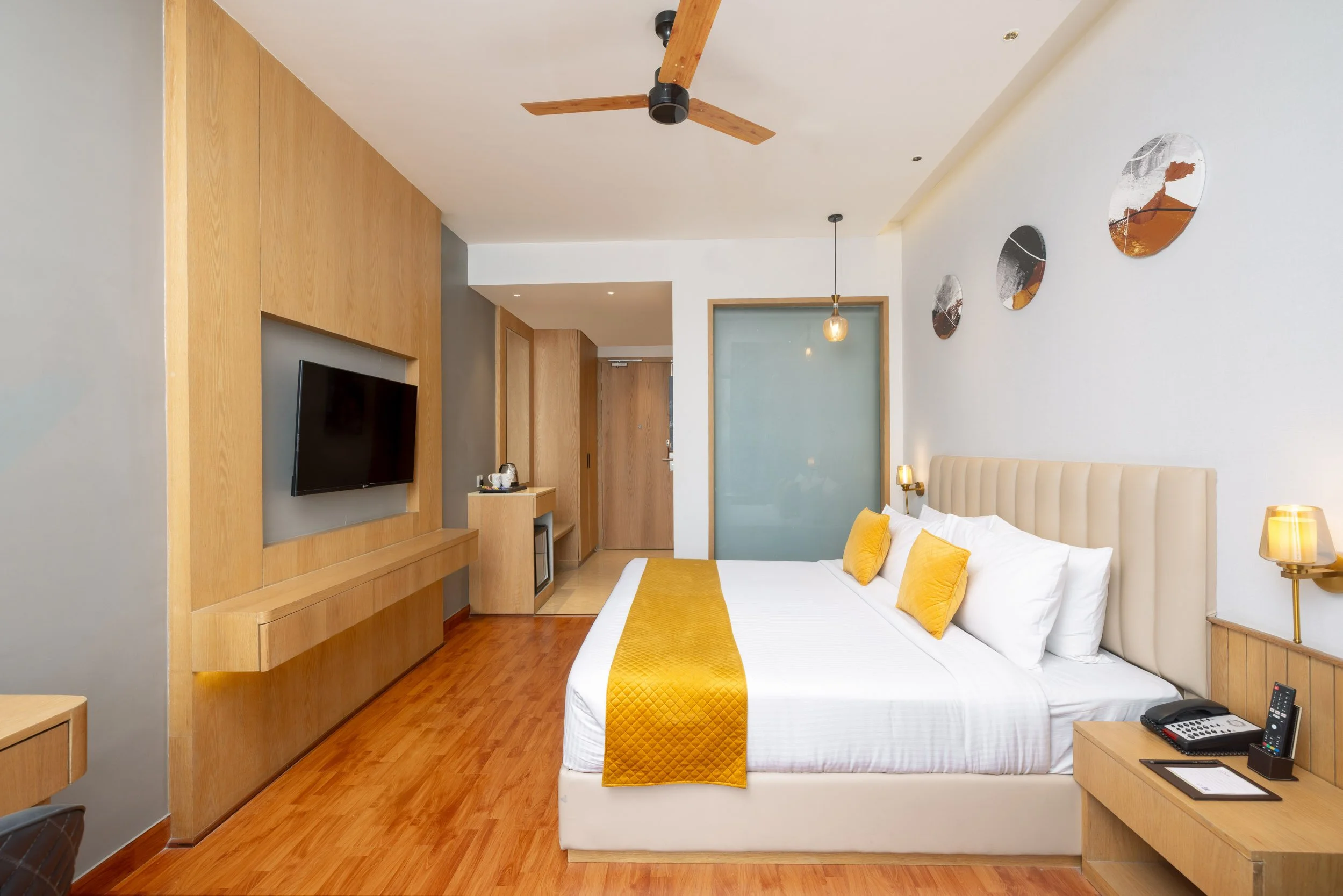 Interior photography of a modern hotel king room featuring a bed with a yellow runner, wood wall paneling, a ceiling fan, and a large window with sheer curtains