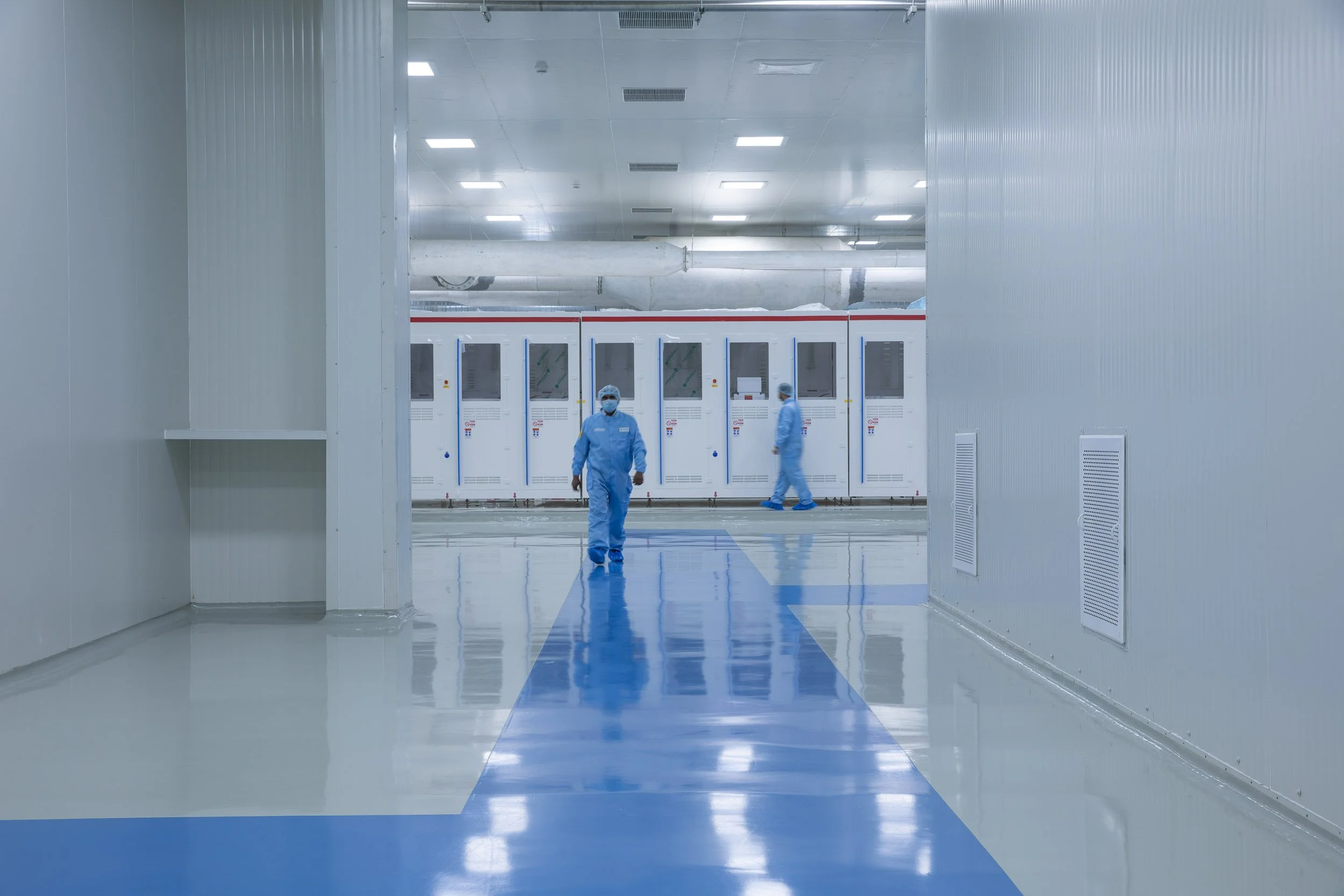 Interior industrial photography of a worker in a blue cleanroom suit walking down a highly sterile, brightly lit corridor in an advanced manufacturing facility.
