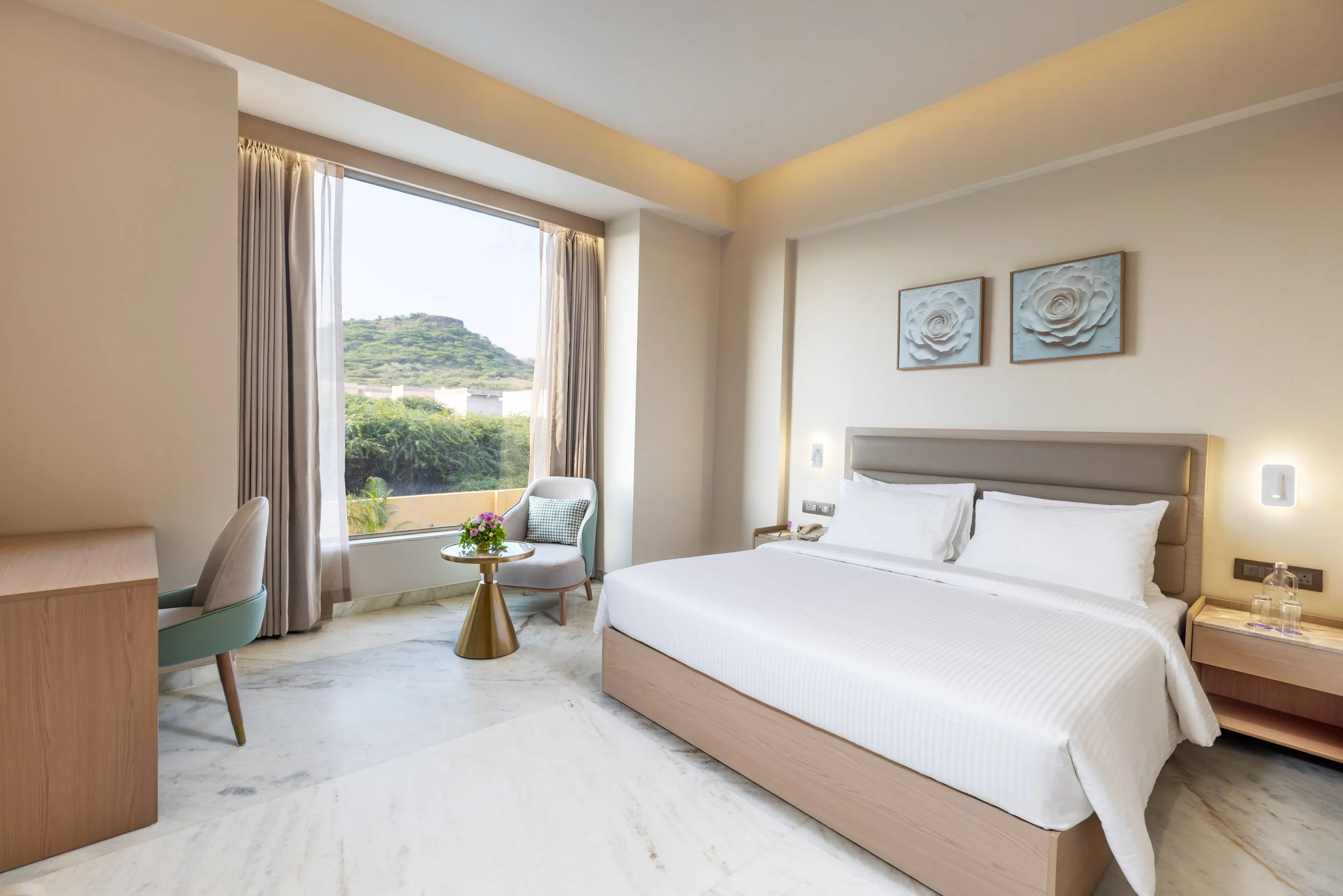 Interior photography of a bright hotel king room featuring a bed with a wood headboard, a seating area with green velvet chairs by a large window, a blue patterned rug, and warm wooden flooring.