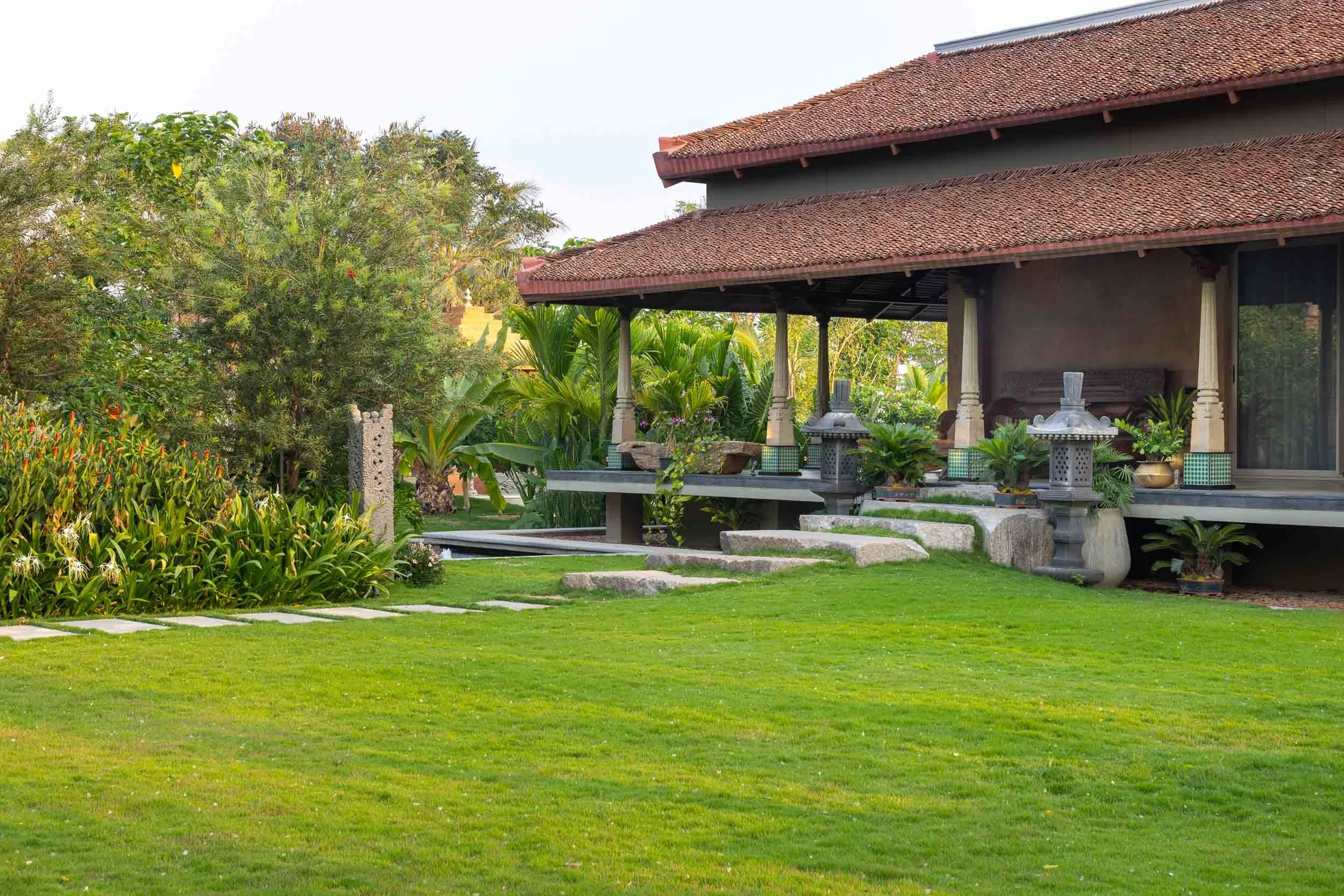 Exterior daytime photography of a luxury resort farm stay featuring a sprawling green lawn, a winding stone pathway, and a traditional building with a covered veranda