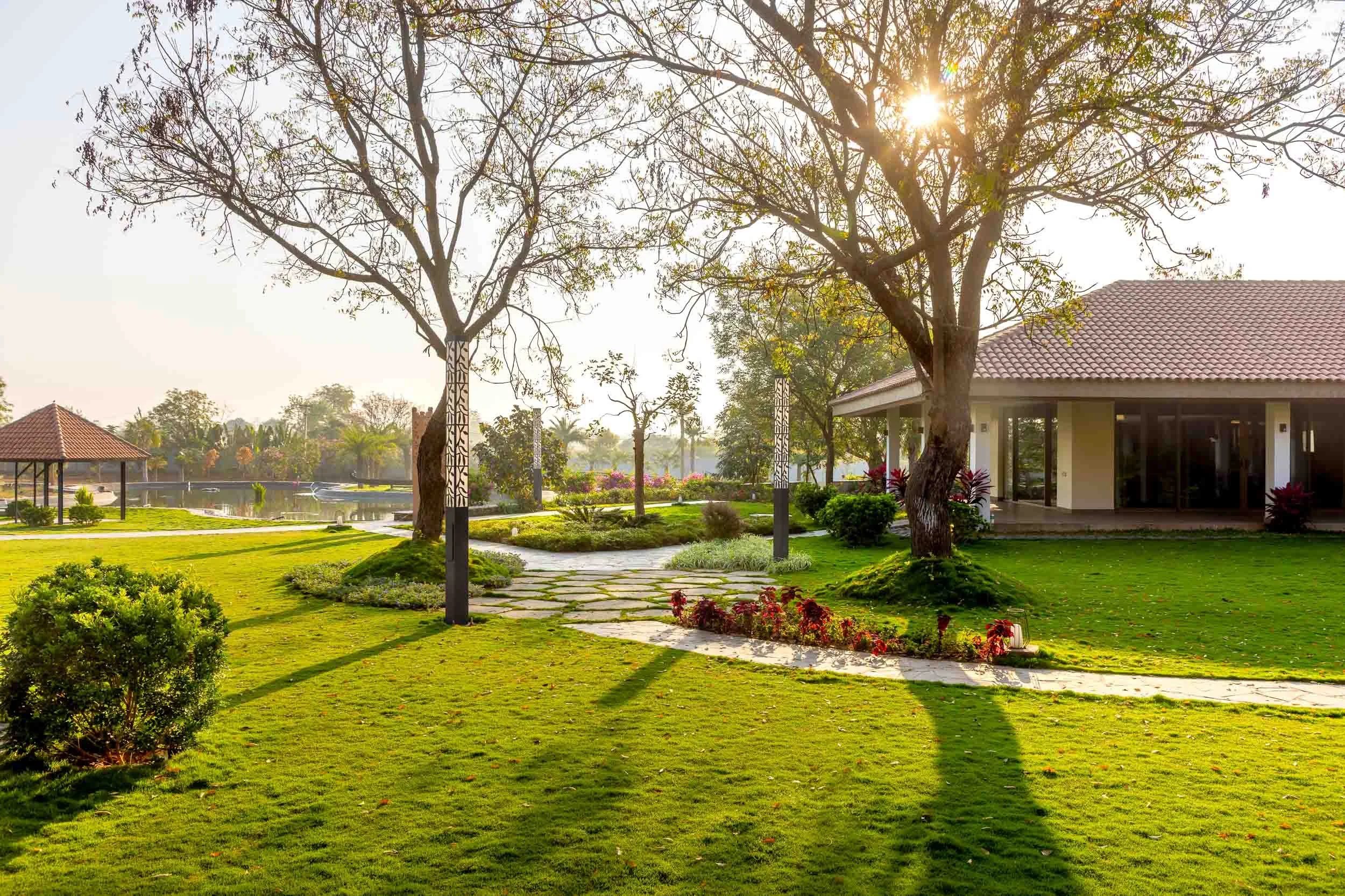 Exterior daytime photography of a luxury resort farm stay featuring a sunlit green lawn, a winding stone pathway, a mature tree, and a traditional pitched-roof building