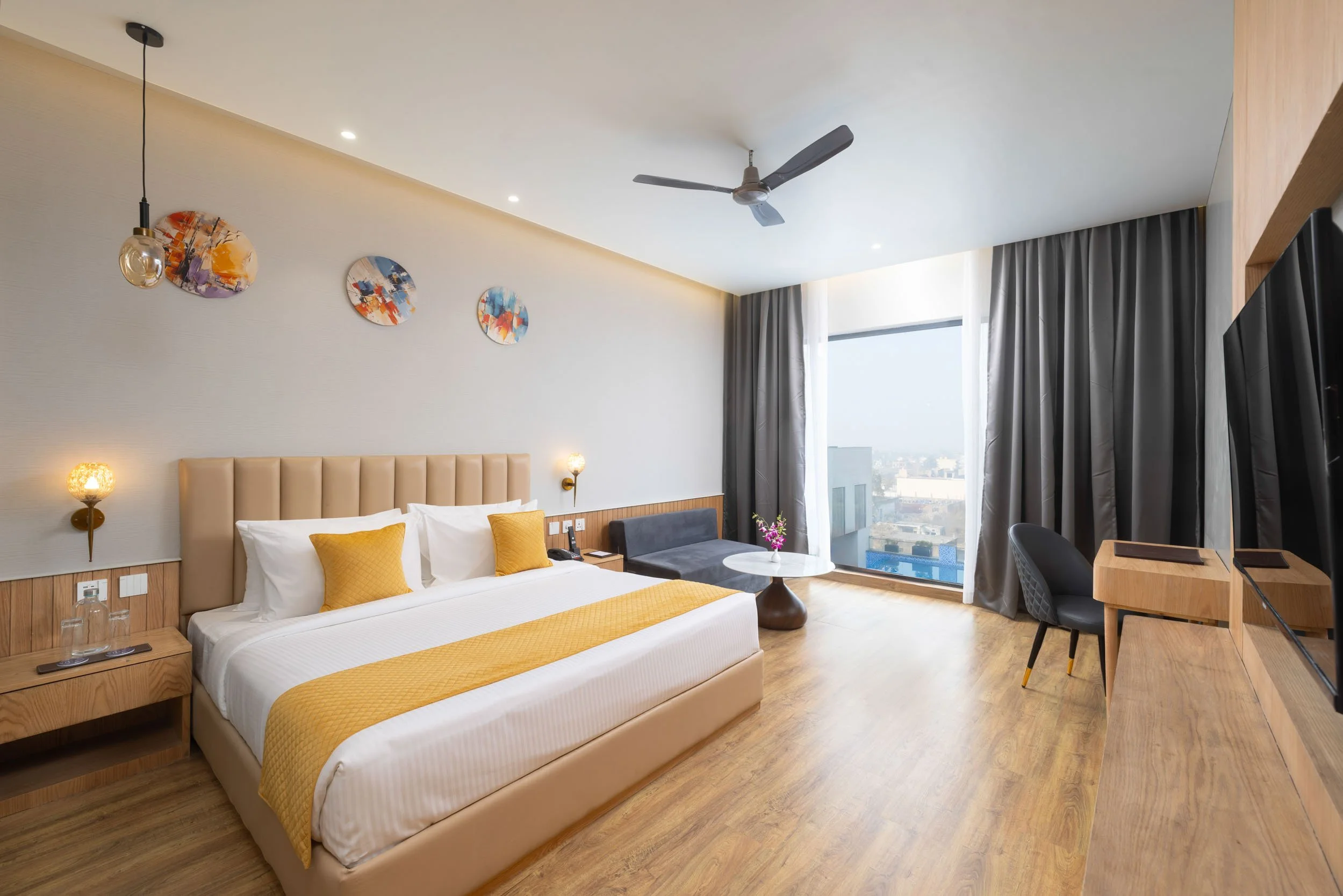 Interior photography of a modern hotel king room featuring a bed with a yellow runner, wood wall paneling, a dark armchair by the window, and a ceiling fan