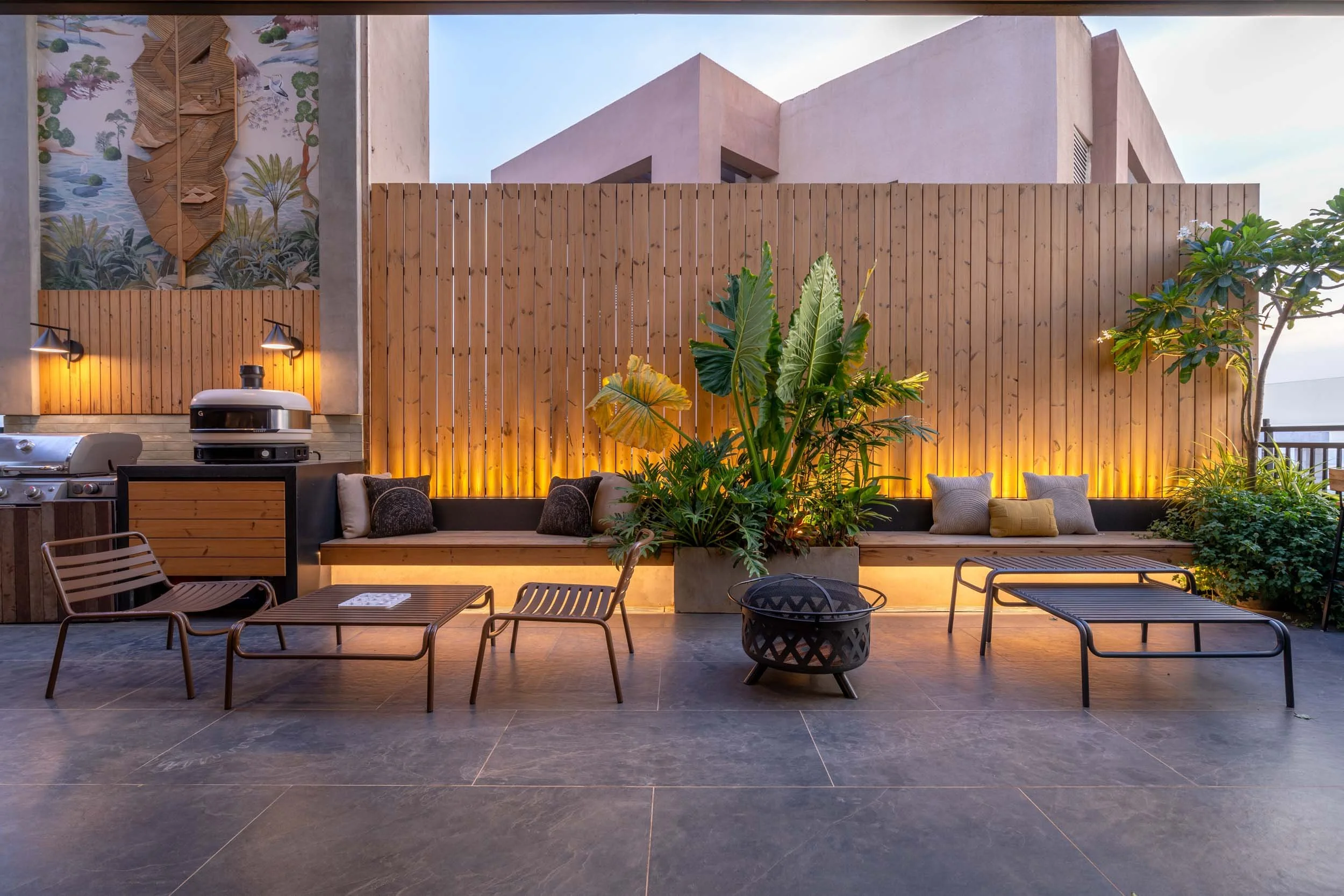 Modern outdoor patio at dusk featuring warm architectural uplighting, a wooden slatted privacy screen, tropical plants, and lounge seating