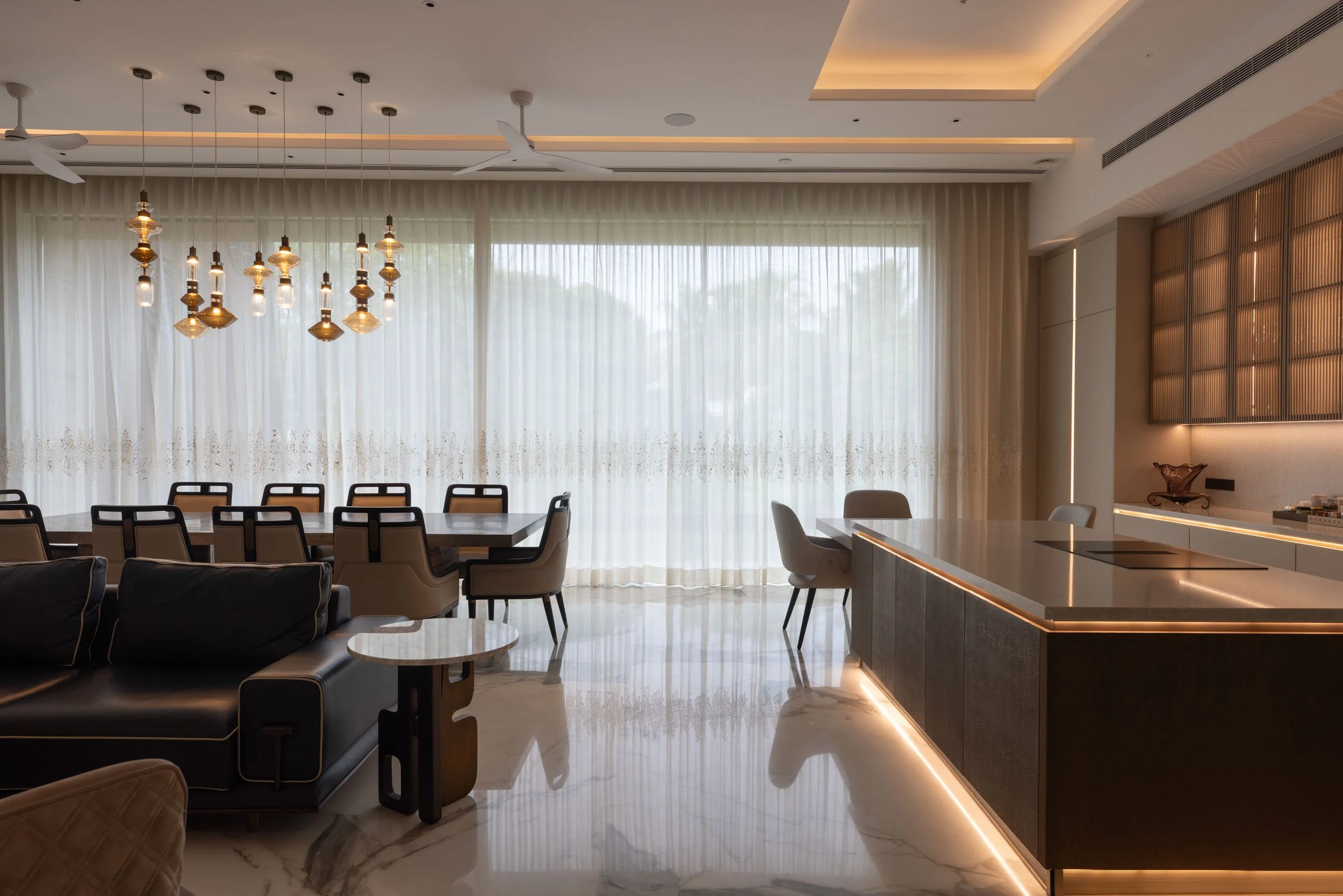 Modern open-plan kitchen and dining area featuring a dark island counter, elegant pendant lighting, and large floor-to-ceiling windows with sheer curtains