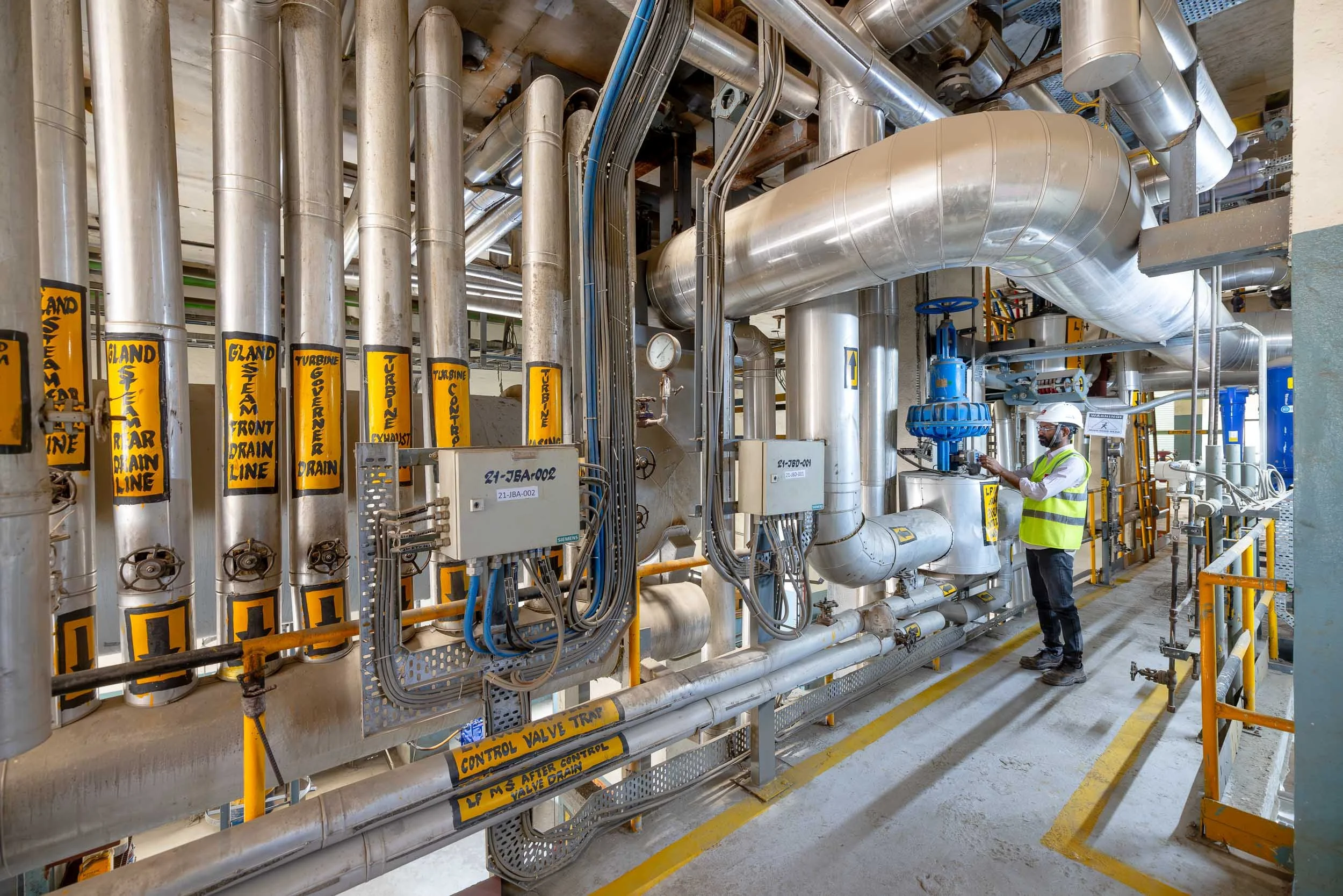 Interior industrial photography of a complex pipe network in a manufacturing plant, featuring large insulated pipes, valves, and a worker in safety gear.