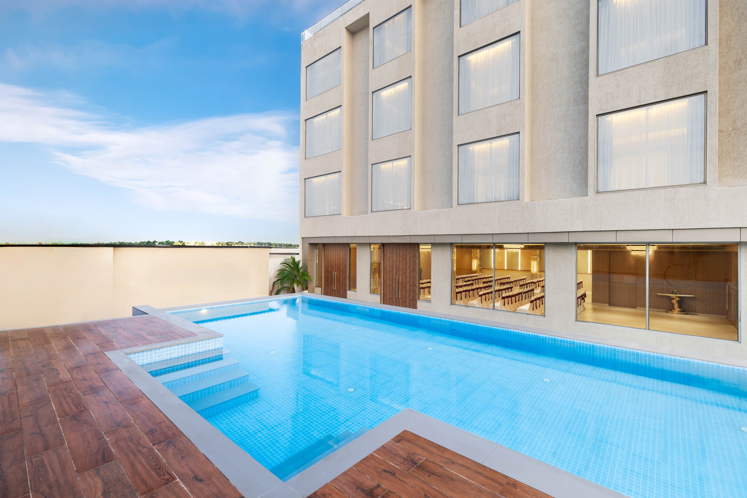 Exterior daytime photography of a modern hotel swimming pool featuring a wooden deck, bright blue water, and a contemporary building facade