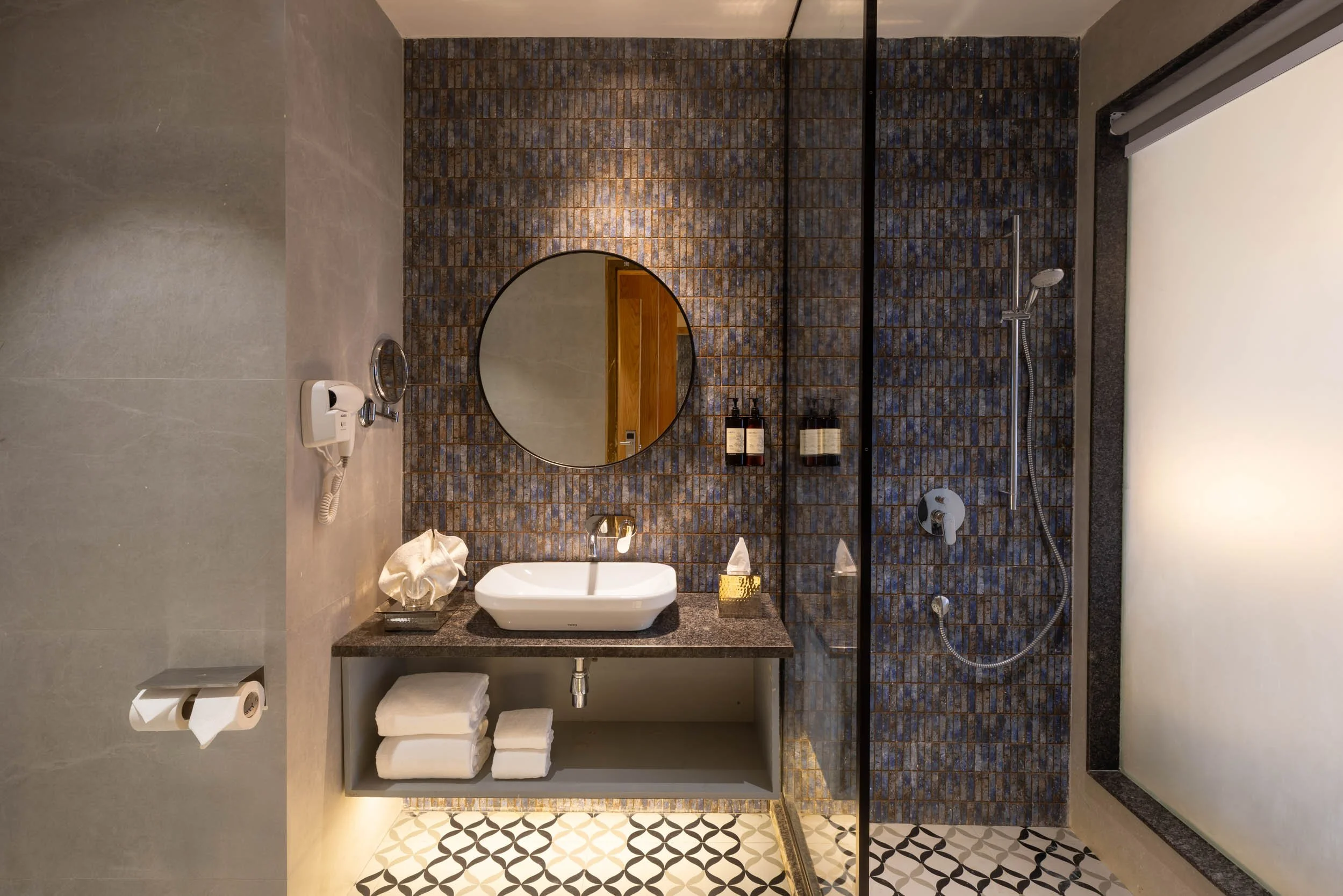 Interior photography of a modern luxury hotel bathroom featuring dark textured walls, a round mirror, a white vessel sink, and a glass-enclosed shower.