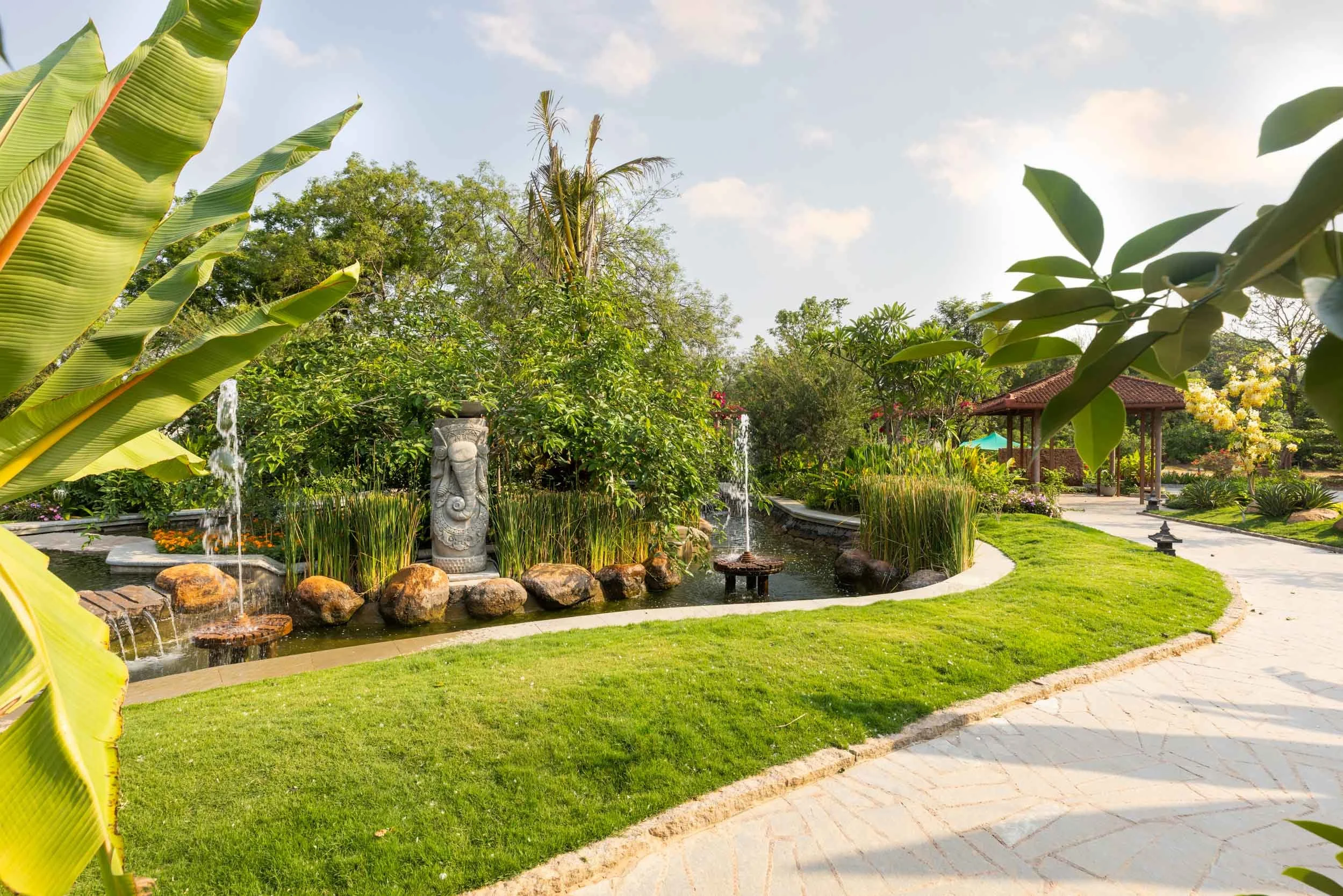 A winding stone pathway meandering through a lush tropical resort garden, featuring banana plants, manicured green lawns, and traditional tiled-roof pavilions