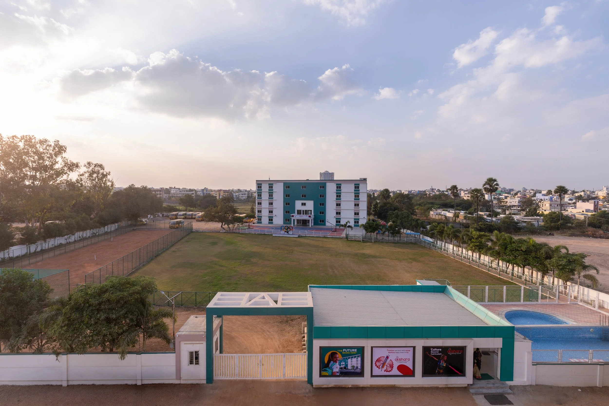 A wide exterior landscape photo of a large, modern, multi-story school building with a sprawling green sports field in the foreground under a cloudy blue sky