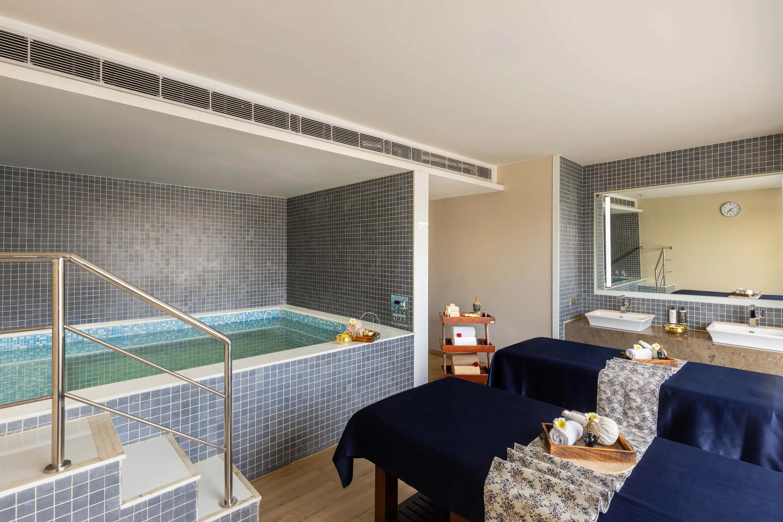 Interior photography of a modern hotel spa wellness room featuring a tiled indoor plunge pool, two massage beds with dark blue towels, and natural light filtering through window blinds