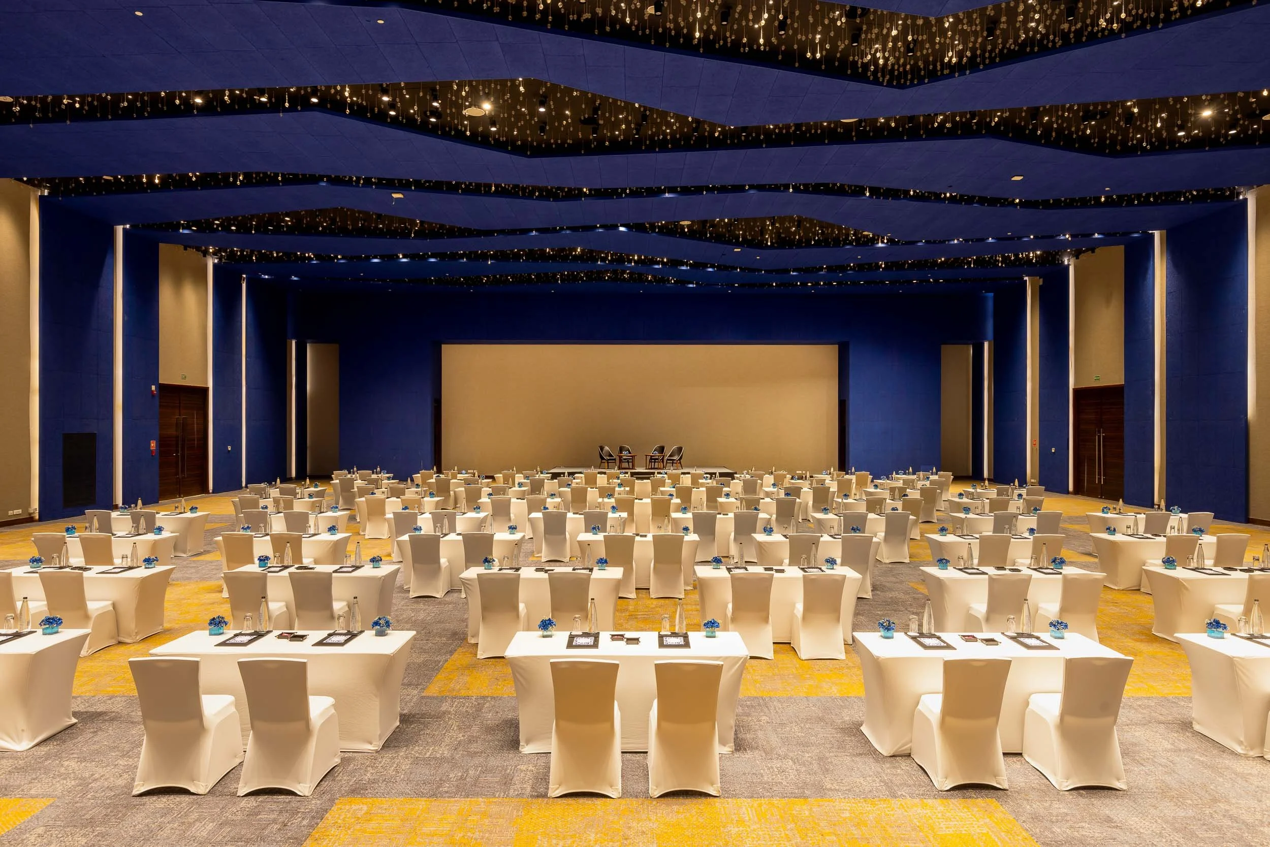 Wide interior photography of a bright hotel banquet hall set up for a large event, featuring long dining tables, gold chairs, deep blue pillars, and a decorative starry ceiling