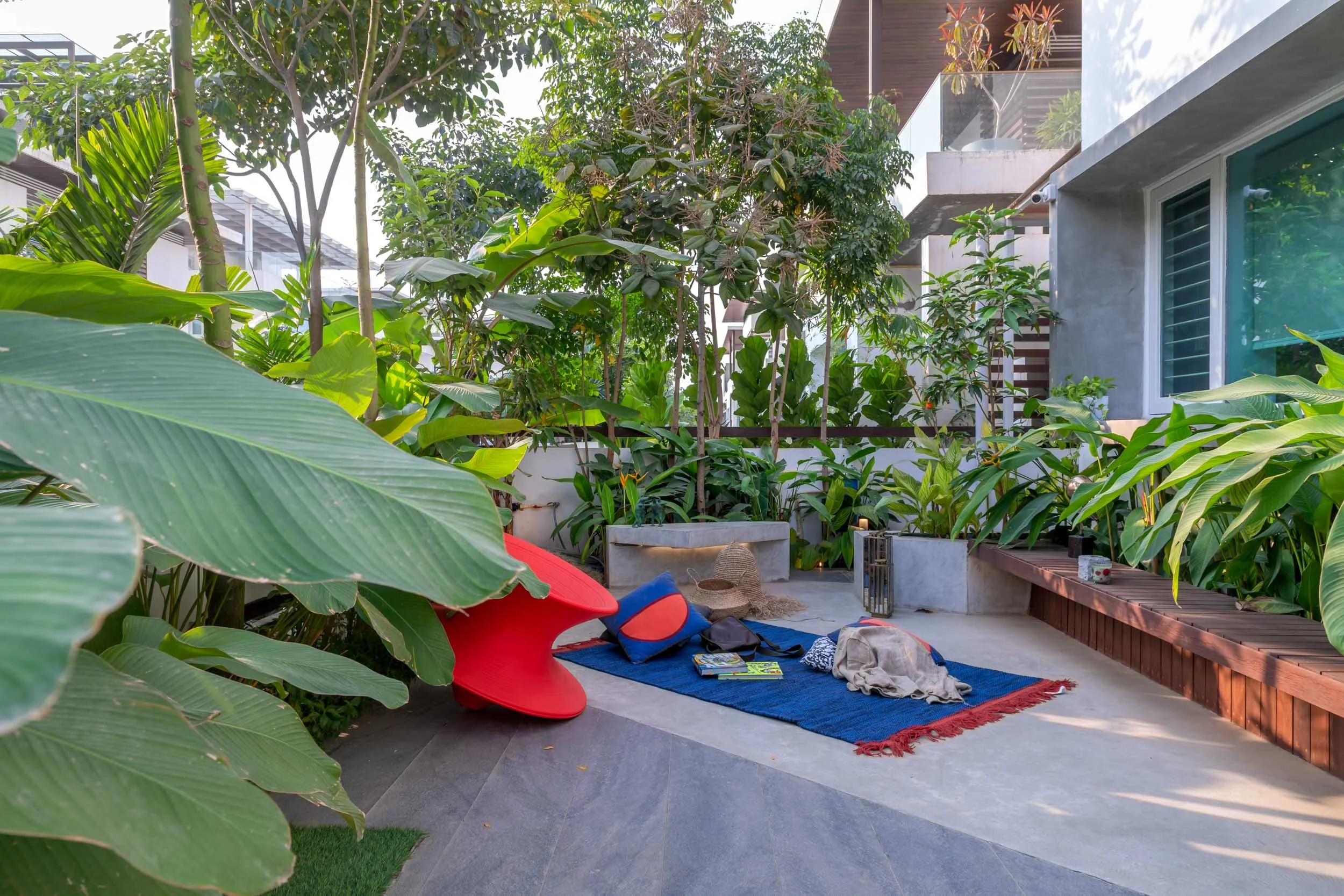 Lush tropical landscape design featuring a wooden deck pathway leading to a modern outdoor seating area with bright red chairs and a blue rug